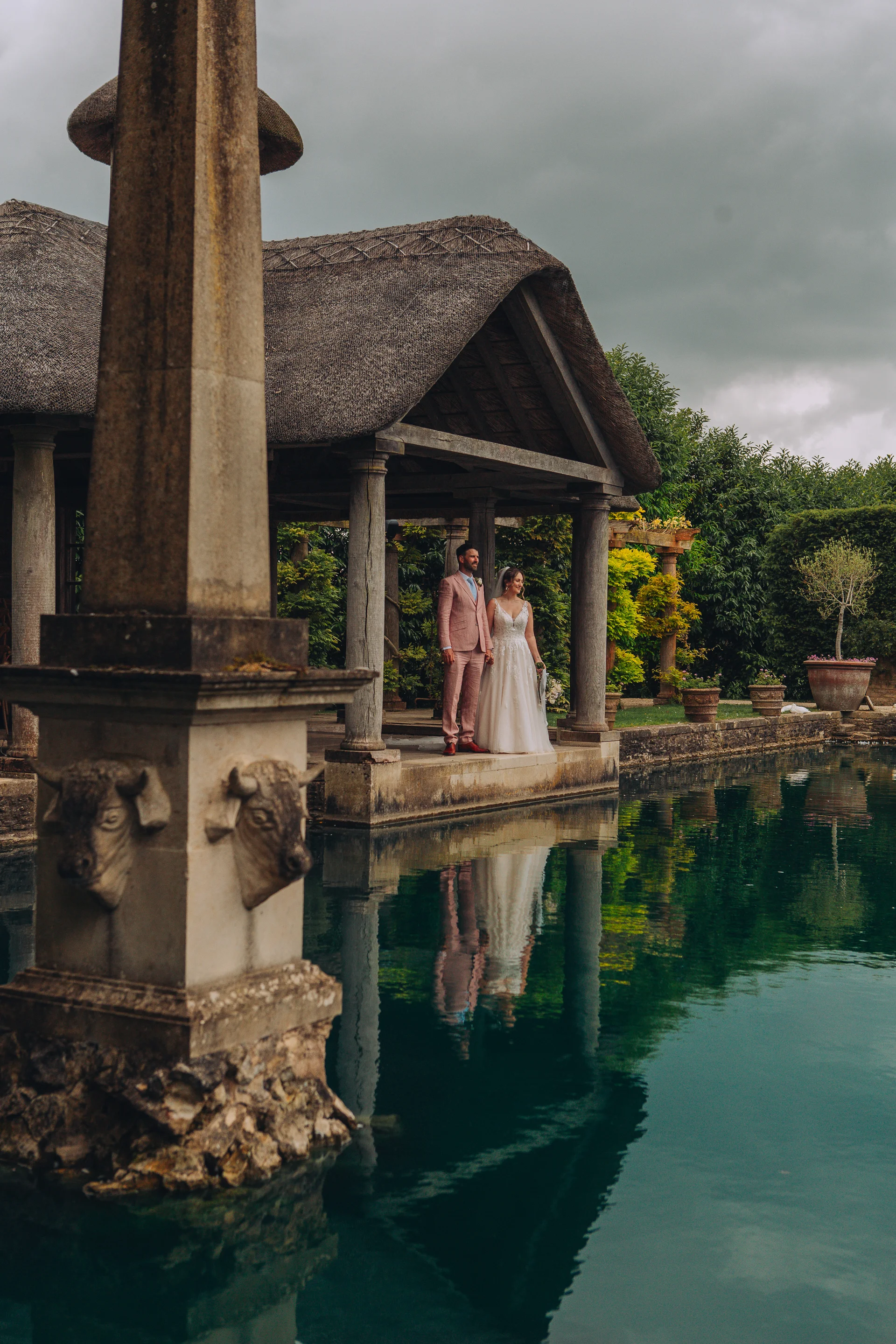 A newlywed couple stands together under a thatched open pavilion by a serene lake, casting a clear reflection in the water beneath an overcast sky.