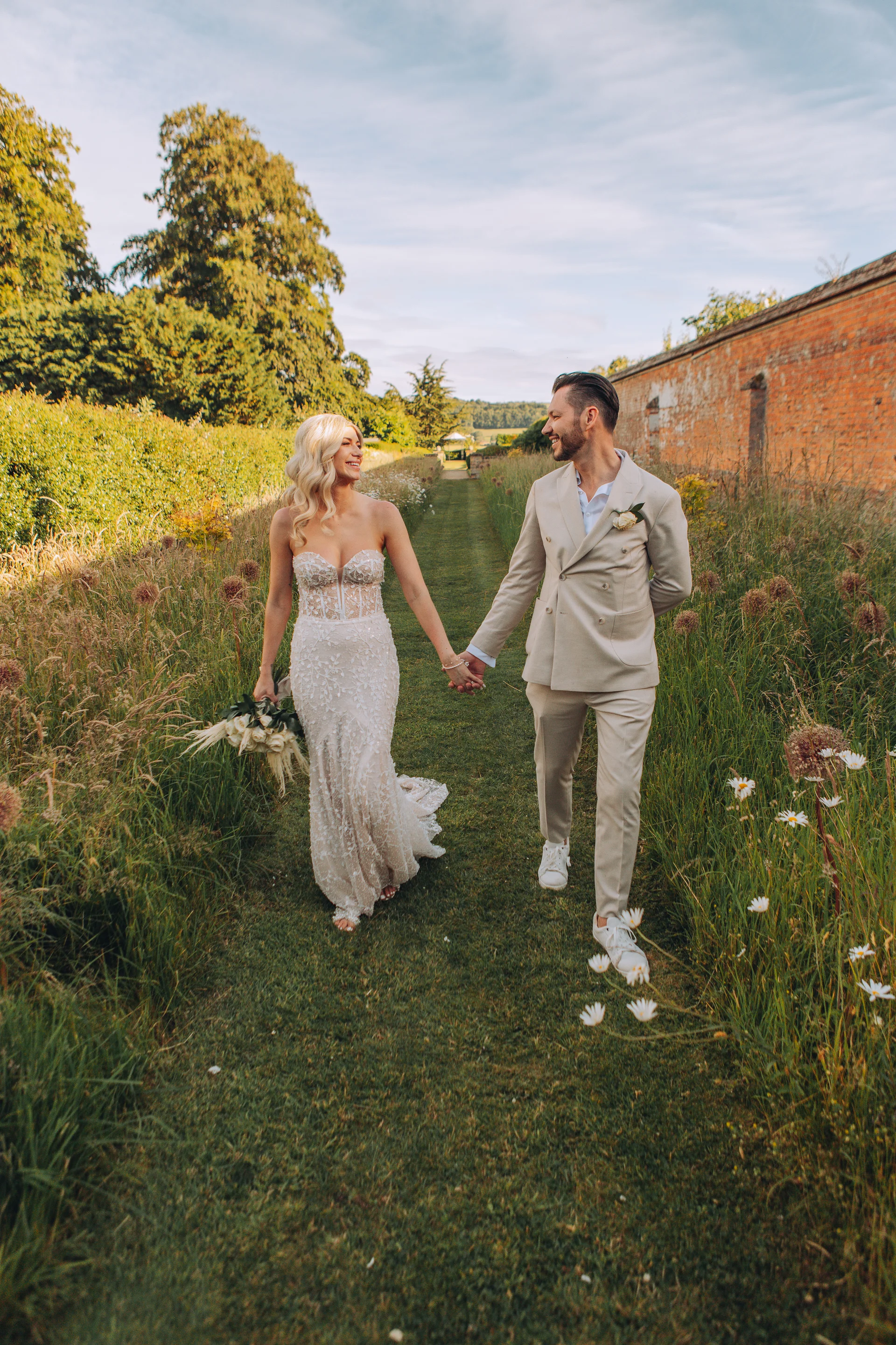 A smiling bride and groom walk hand in hand down a grassy path flanked by wildflowers and an old brick wall during golden hour.