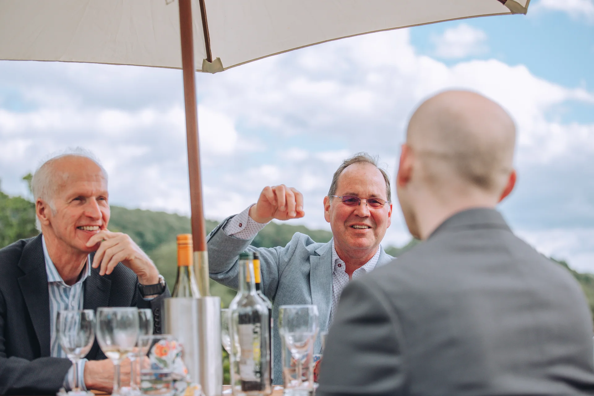 Guests are captured laughing and talking outdoors under the shade of a parasol during a wedding celebration.