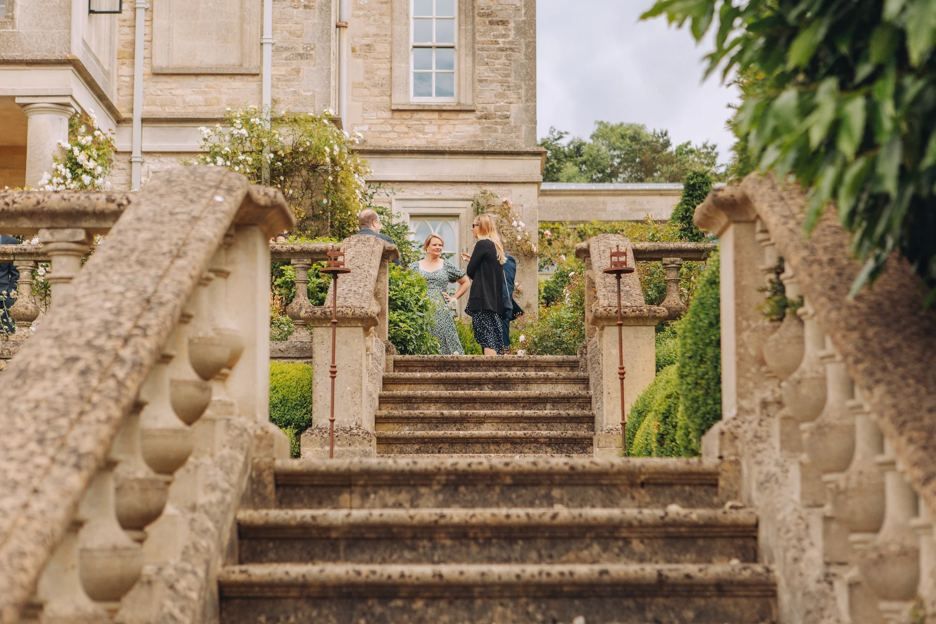 Wedding guests mingle at the top of an ornate stone staircase framed by lush greenery and a classic country house exterior.