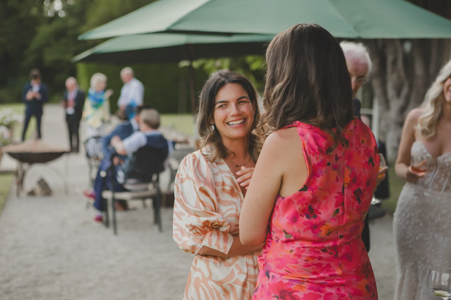 Wedding guests in colourful patterned dresses laugh and chat together during an outdoor drinks reception beneath green parasols.