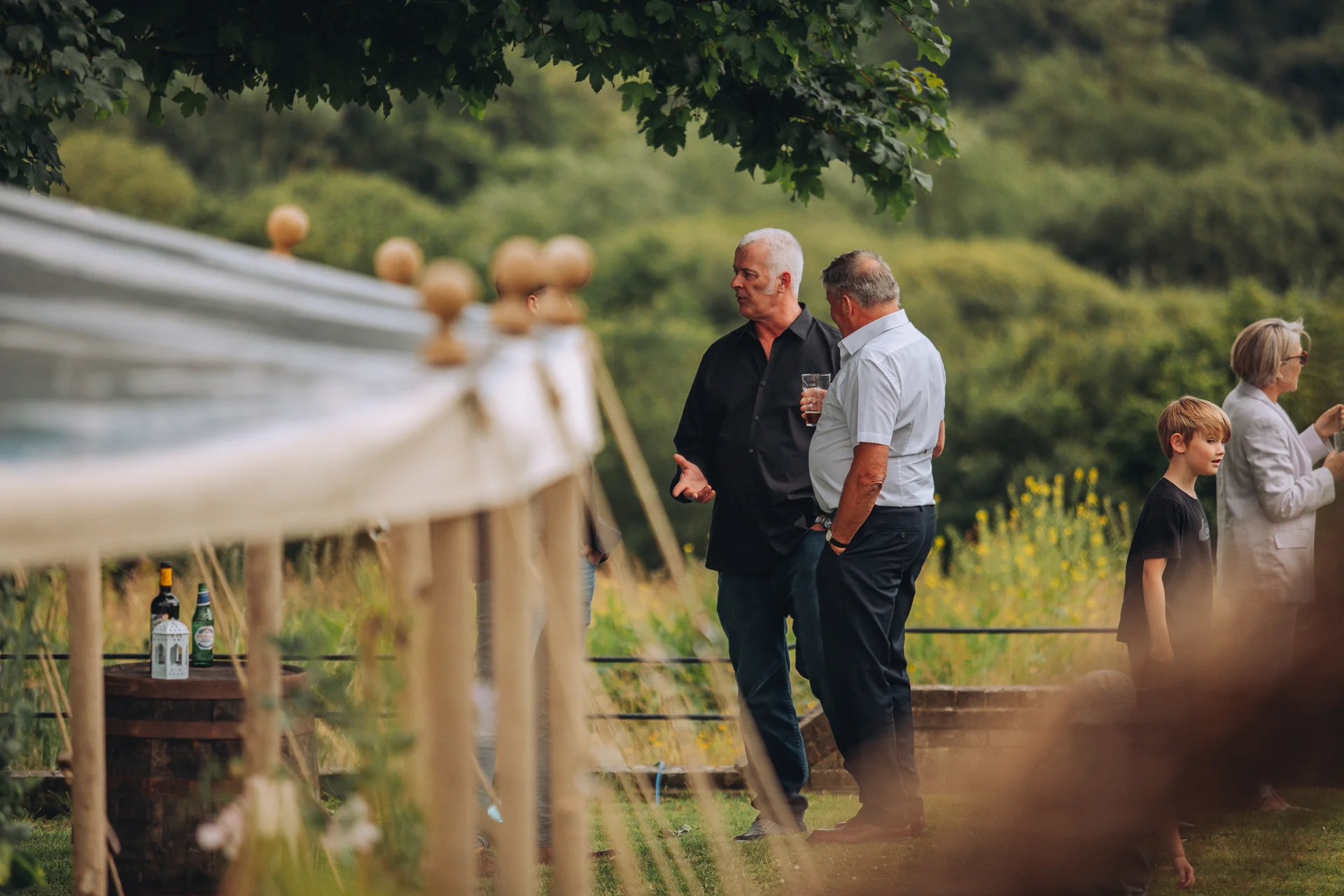 Wedding guests mingle outdoors near a marquee during a relaxed summer drinks reception, with children and family members chatting in the sunlight.