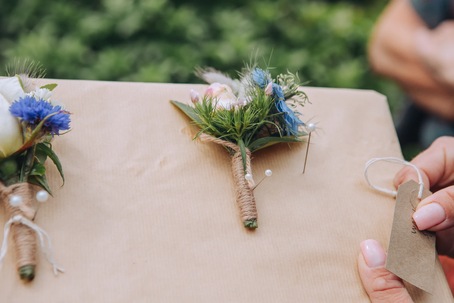 Two handmade rustic buttonholes tied with twine and featuring blue cornflowers rest on a brown paper package held by hands with pink manicured nails.