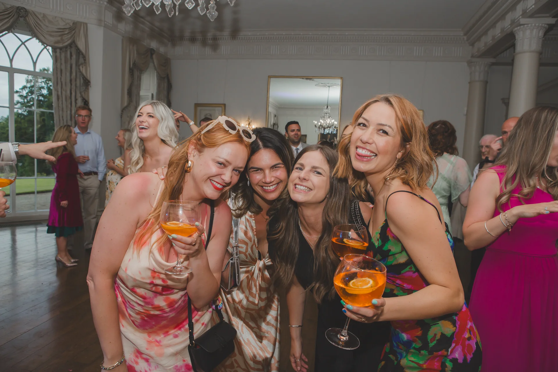 A group of wedding guests enjoying drinks and laughing together at a reception inside a grand ballroom.