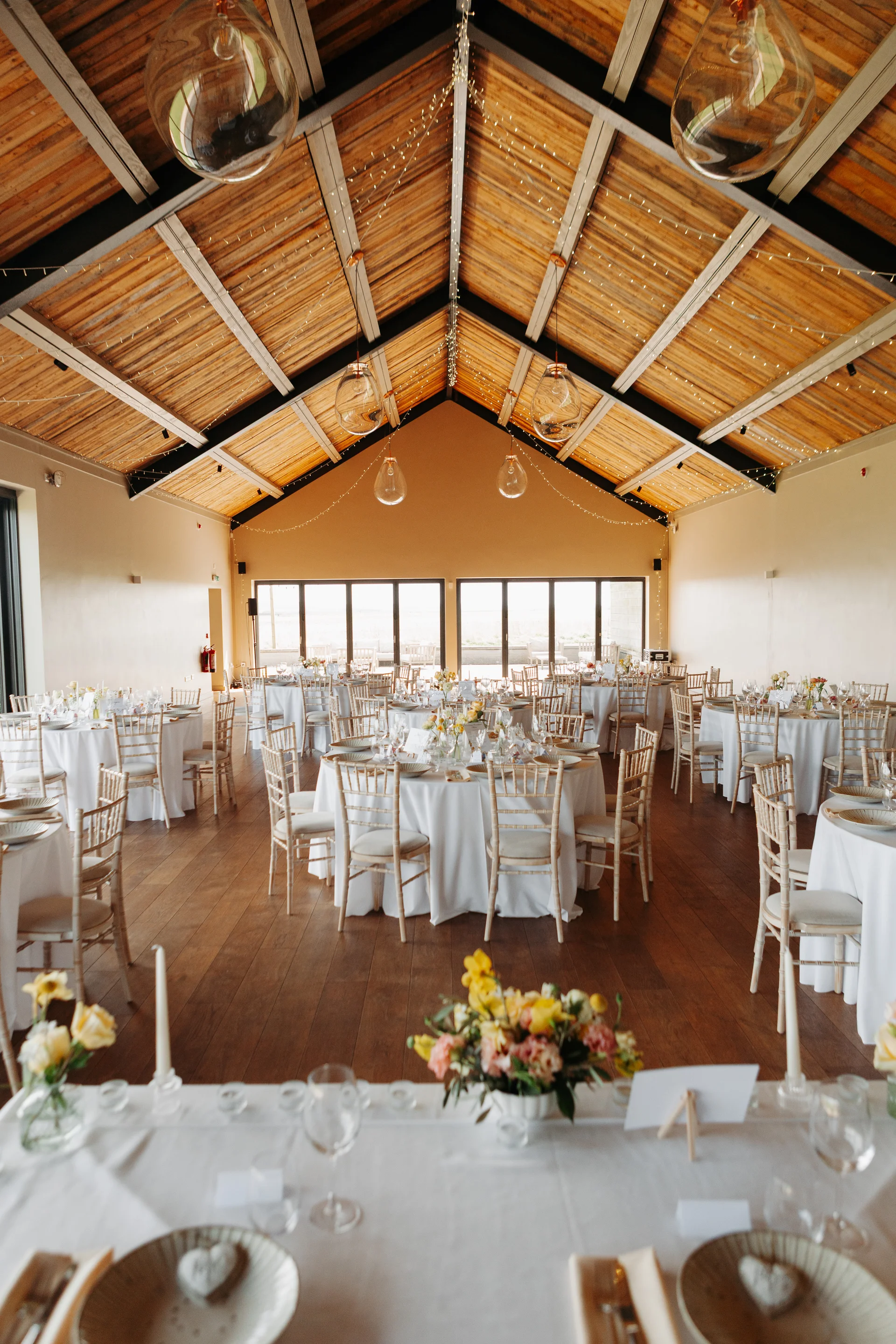 An empty wedding reception venue featuring a vaulted wooden ceiling, exposed beams, round tables with white linens, and large glass pendant lights.