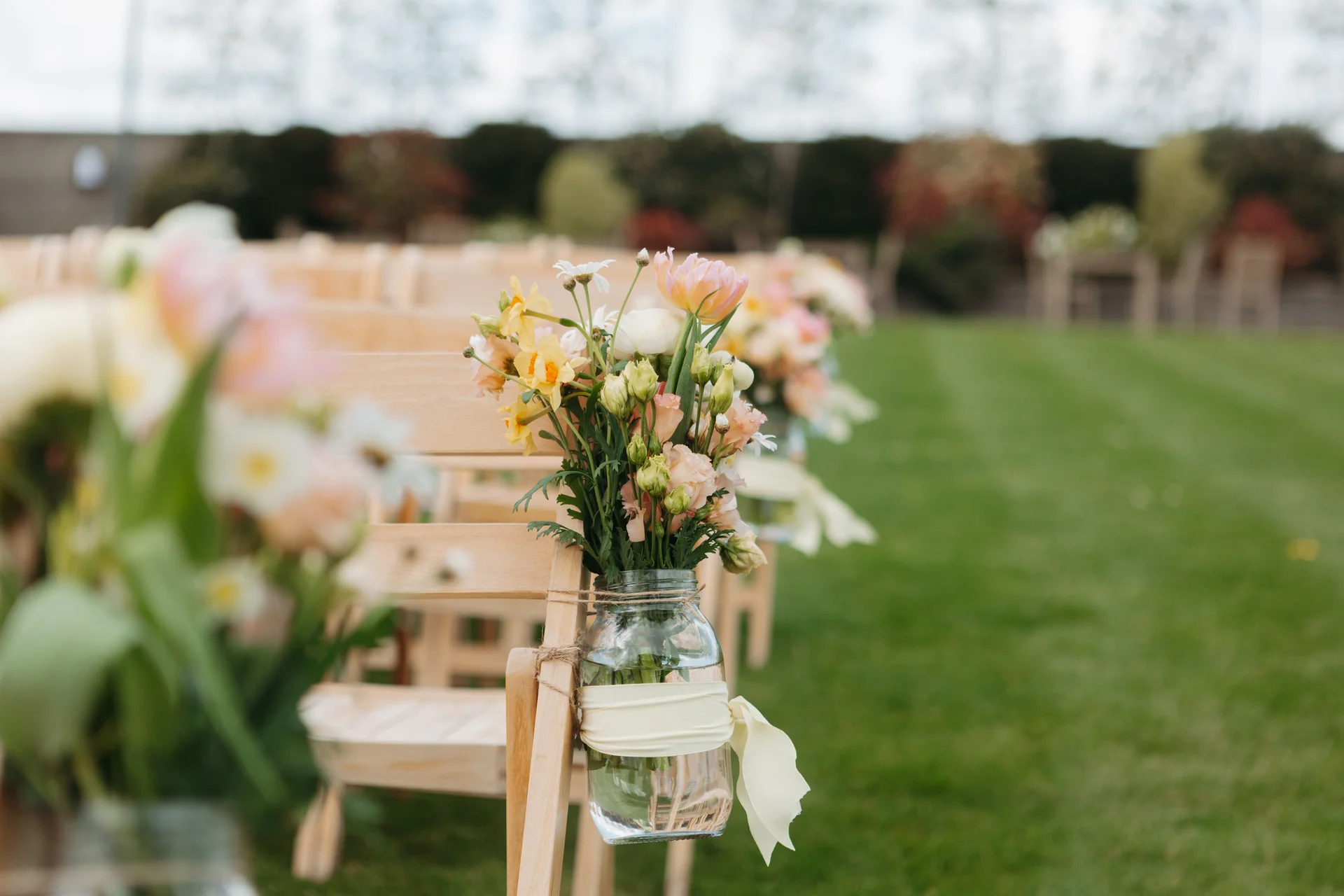A row of wooden folding chairs set up for an outdoor ceremony features mason jars tied with twine and ribbon, filled with colourful pastel spring flowers.