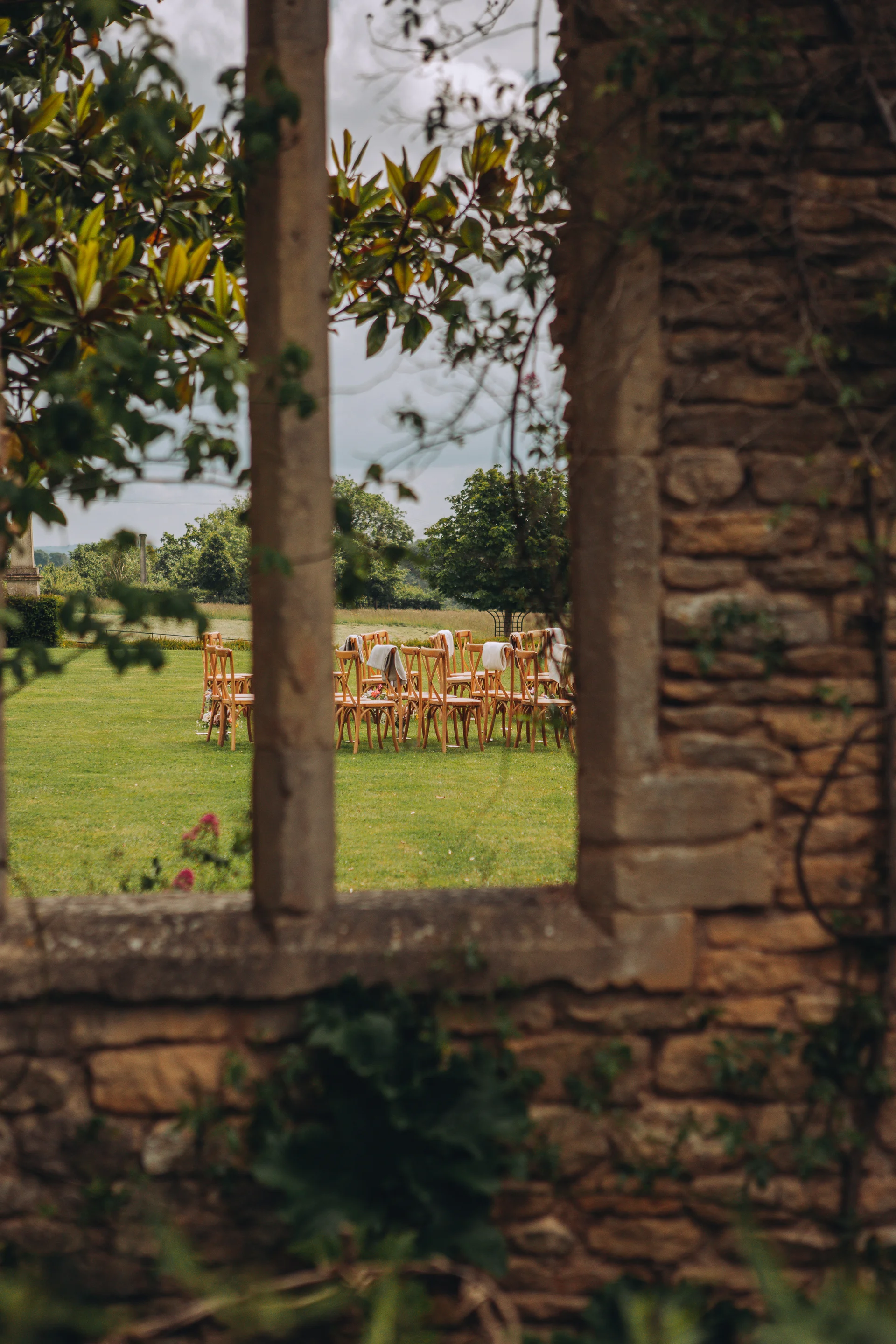 An outdoor wedding ceremony setup in the round on a grassy lawn is framed through an old stone architectural archway and rustic wall.