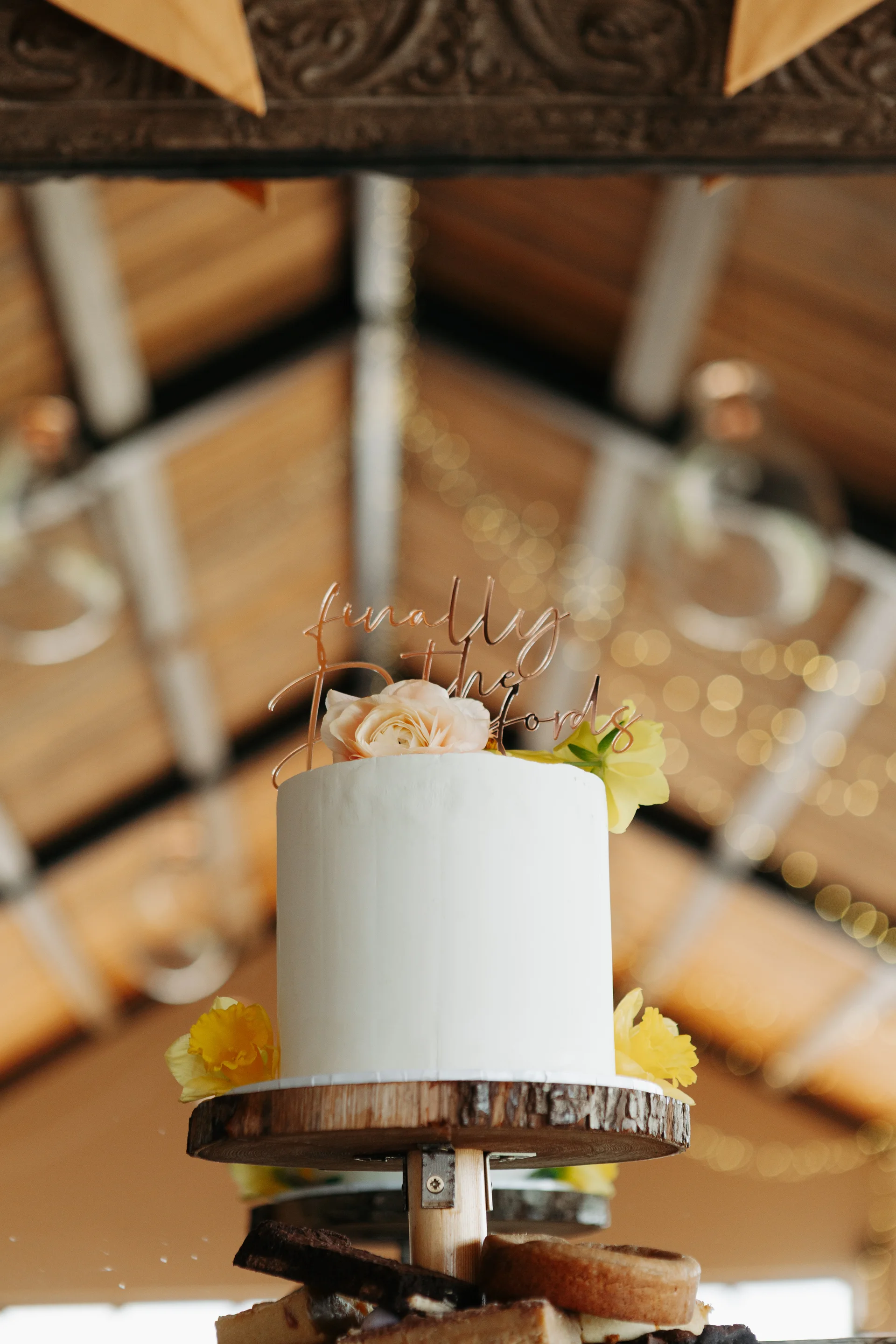 A single-tier white buttercream wedding cake is displayed on a rustic wood slice stand inside a barn with exposed rafters and fairy lights.