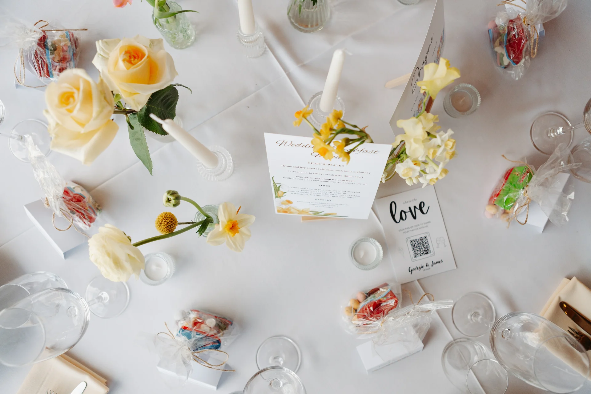 An overhead view of a wedding reception tablescape featuring butter yellow roses, bud vases, taper candles, menu cards, and individual guest favours on a white tablecloth.