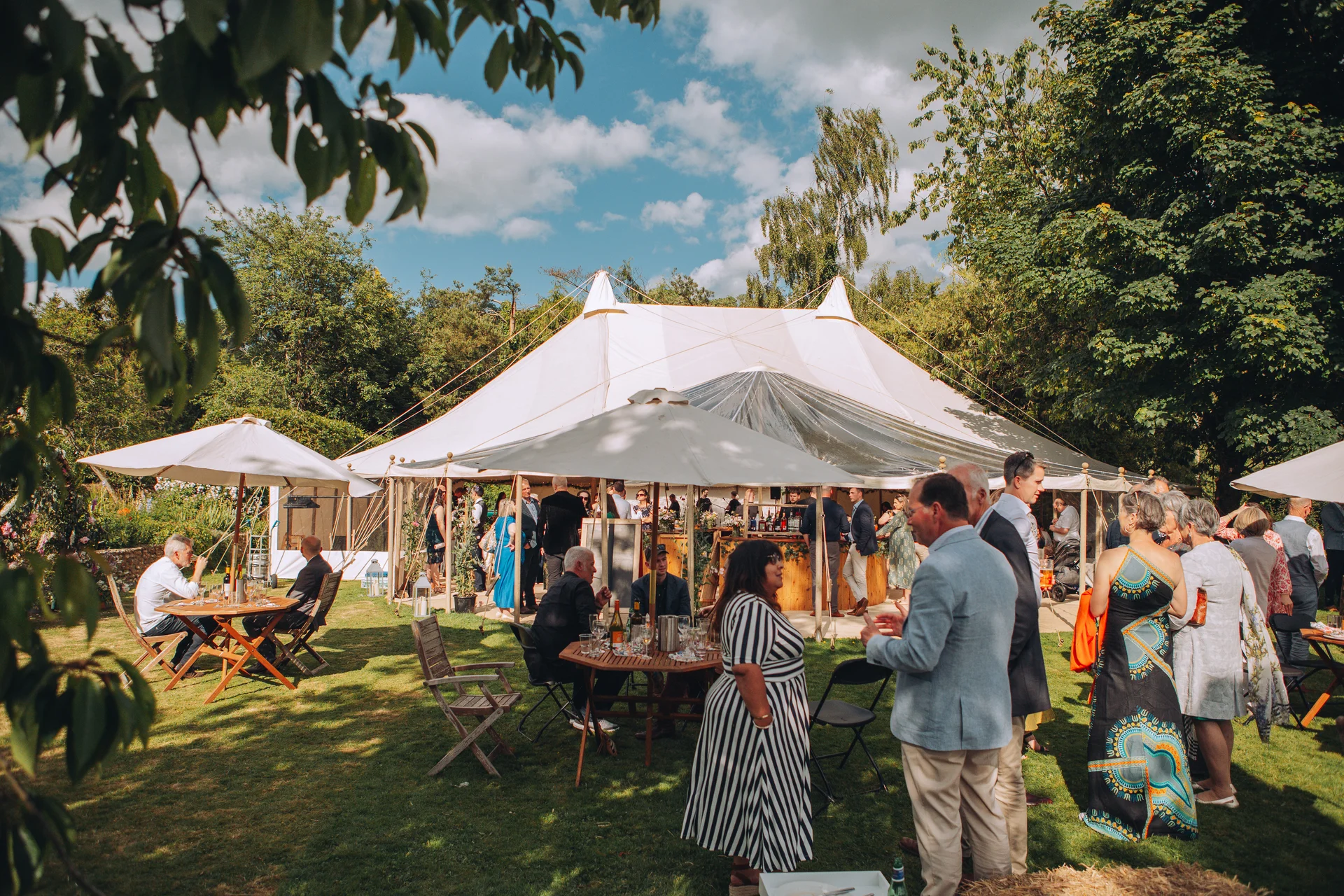 Wedding guests mingle outside a large sailcloth marquee set up on a sunny lawn during an outdoor summer reception.