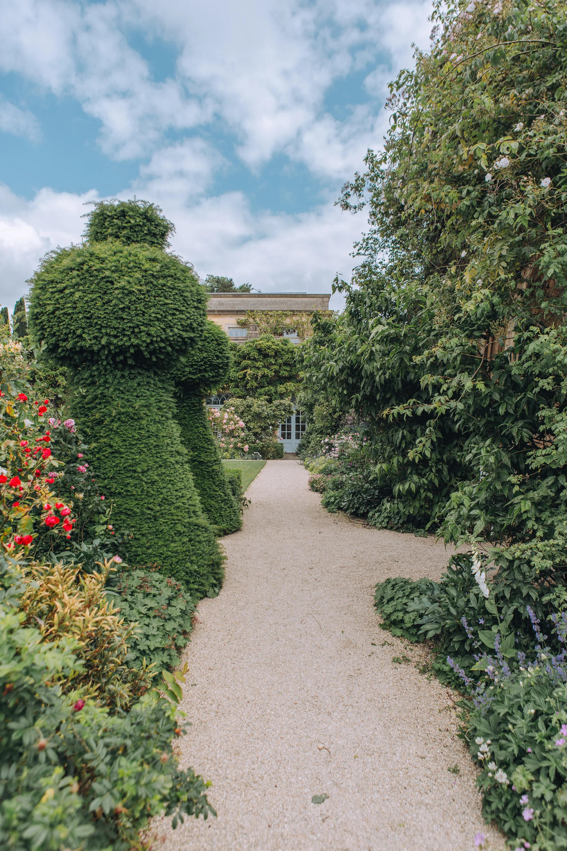 A charming gravel pathway winds through lush manicured gardens towards a stately manor house framed by ornate topiary and climbing roses.