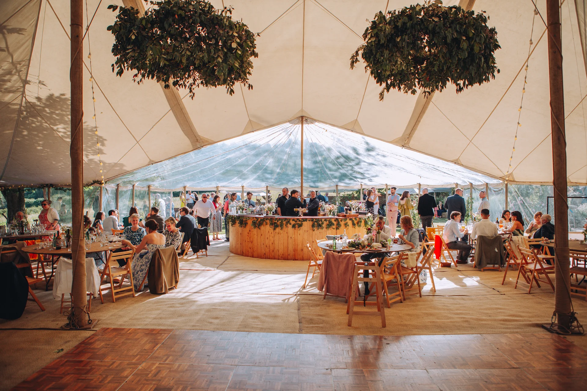Guests mingle and dine inside a spacious sailcloth marquee wedding reception featuring a central wooden circular bar and hanging floral installations.