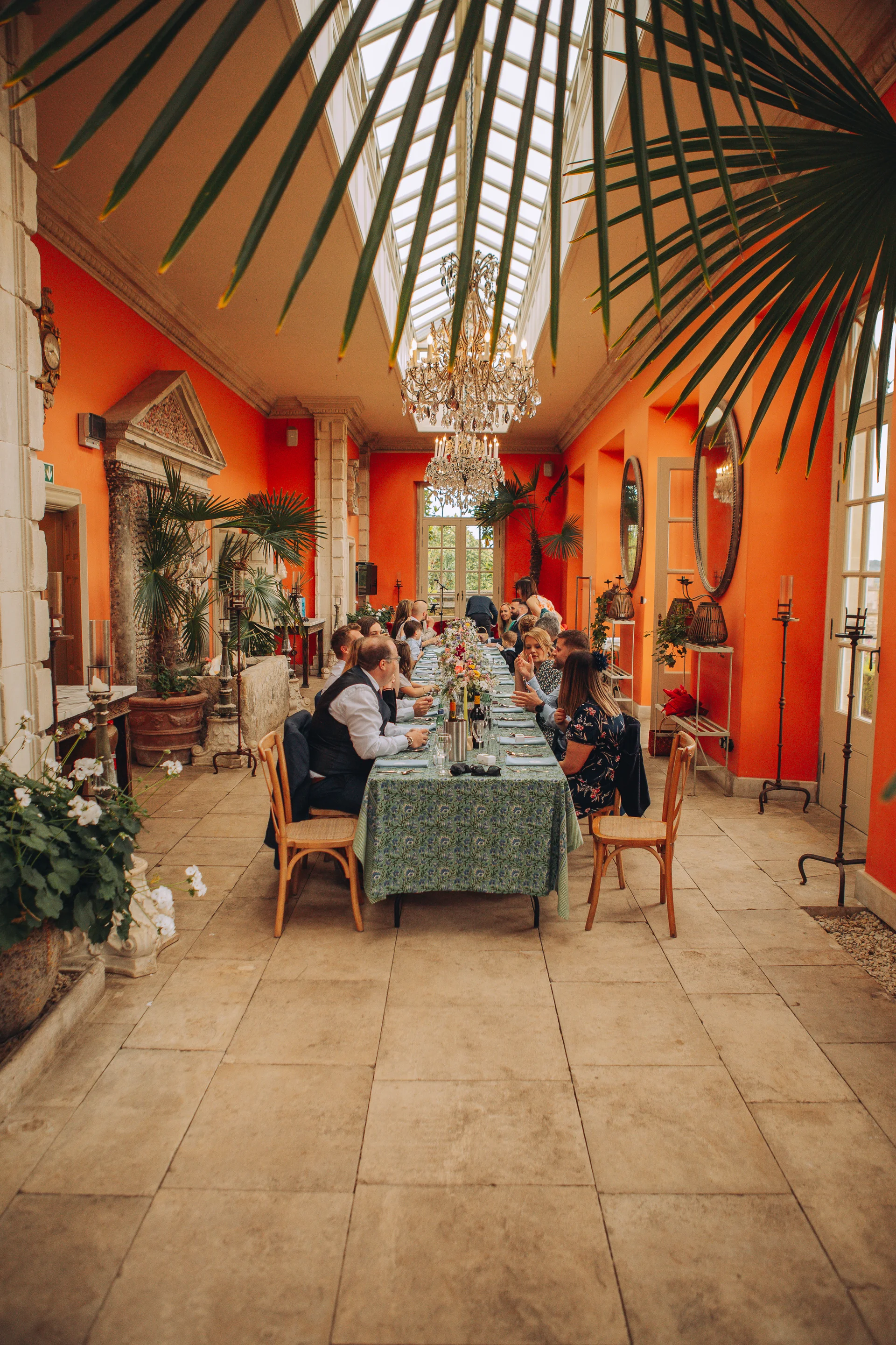 Guests enjoy a wedding breakfast seated at a long banquet table inside a bright orangery featuring vibrant orange walls, a glass ceiling, and crystal chandeliers.