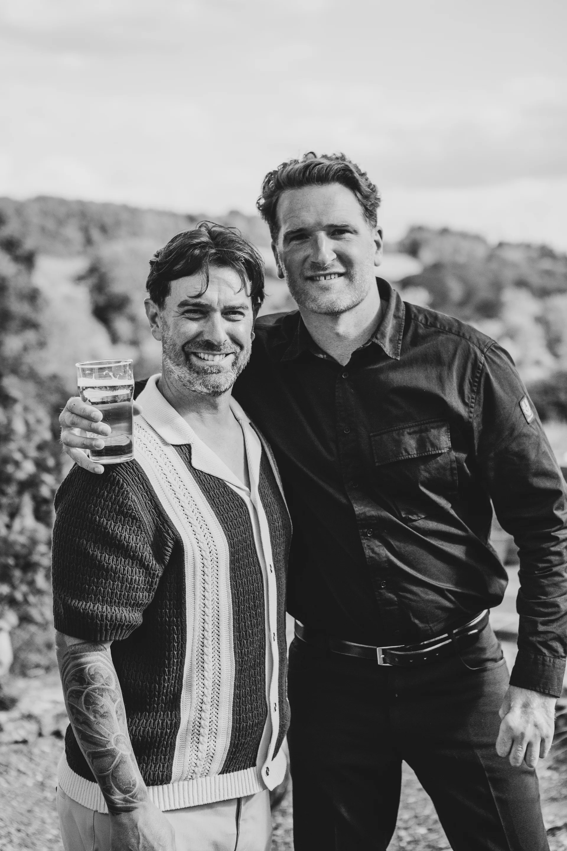 Two smiling grooms pose together outdoors in a relaxed black and white portrait, one holding a drink and wearing a patterned short-sleeved shirt.