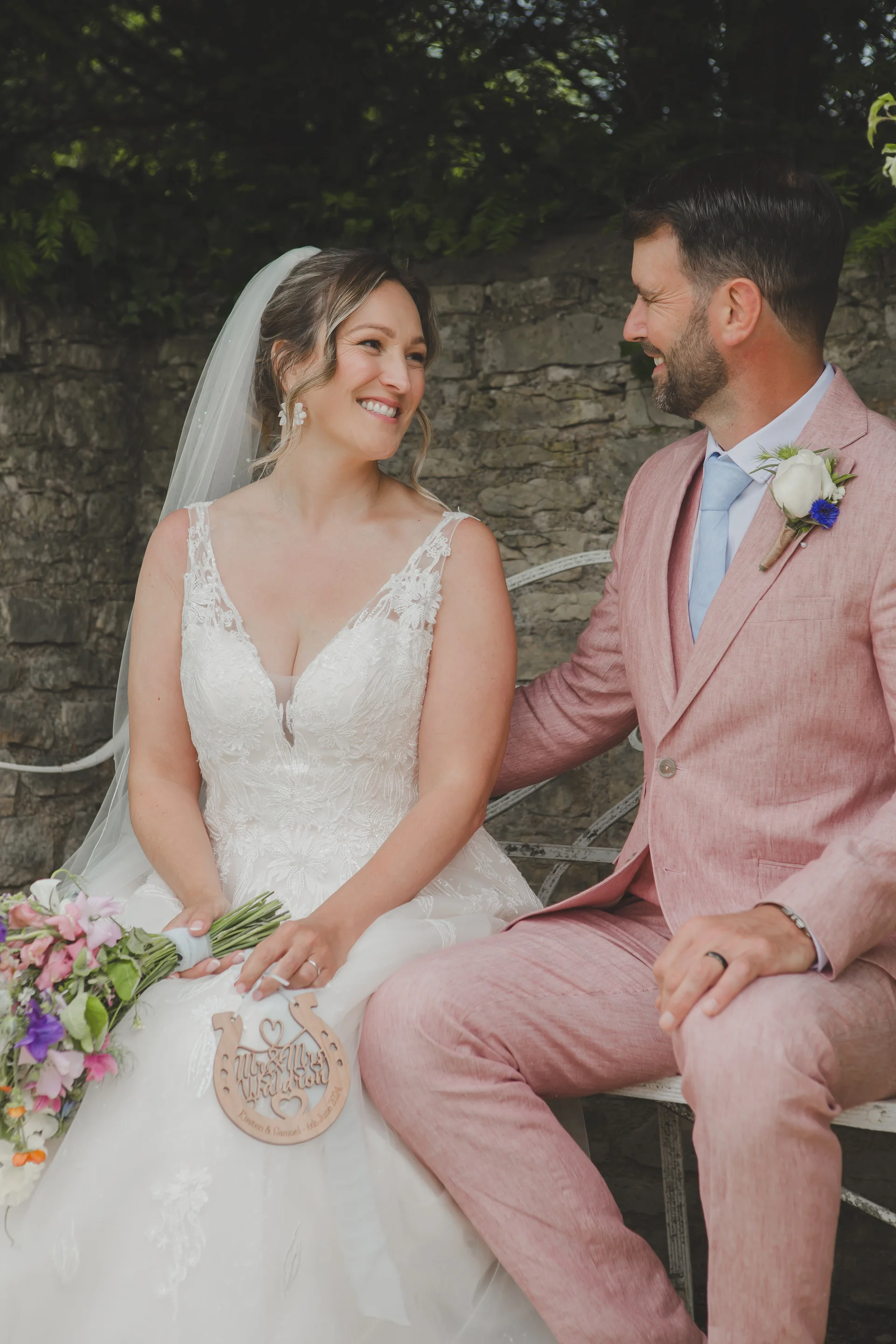 A smiling bride and groom sit together on a white bench against a rustic stone wall, with the bride holding a bouquet and a wooden horseshoe prop.