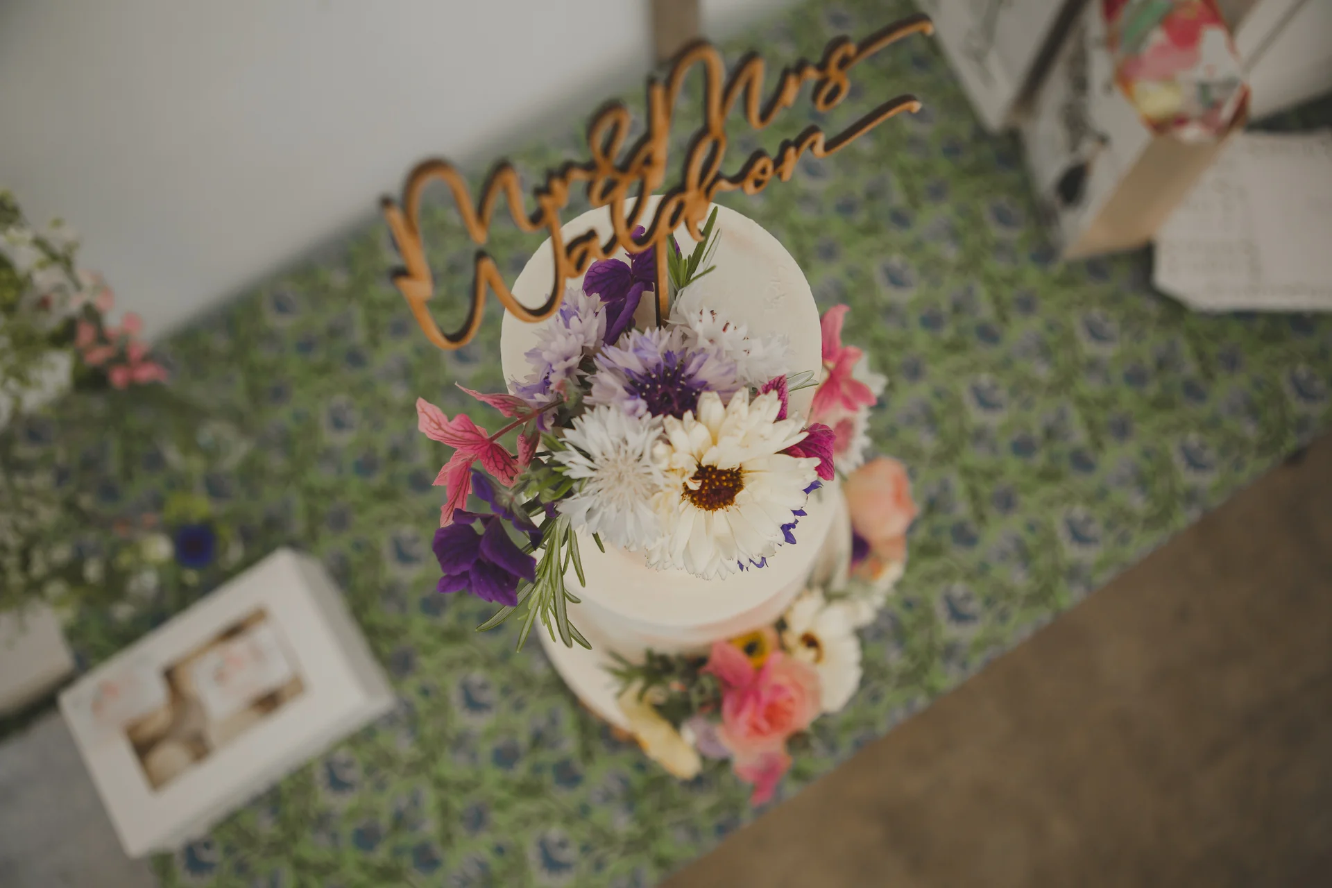 An overhead view of a two-tiered wedding cake adorned with vibrant white, purple, and pink flowers and topped with a wooden script cake topper.