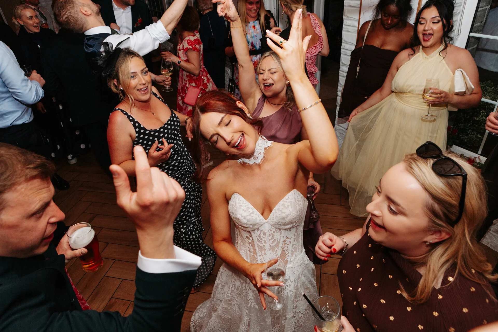 A lively group of wedding guests and the bride in a lace gown with a sweetheart neckline dance enthusiastically on a crowded dance floor under direct flash lighting.