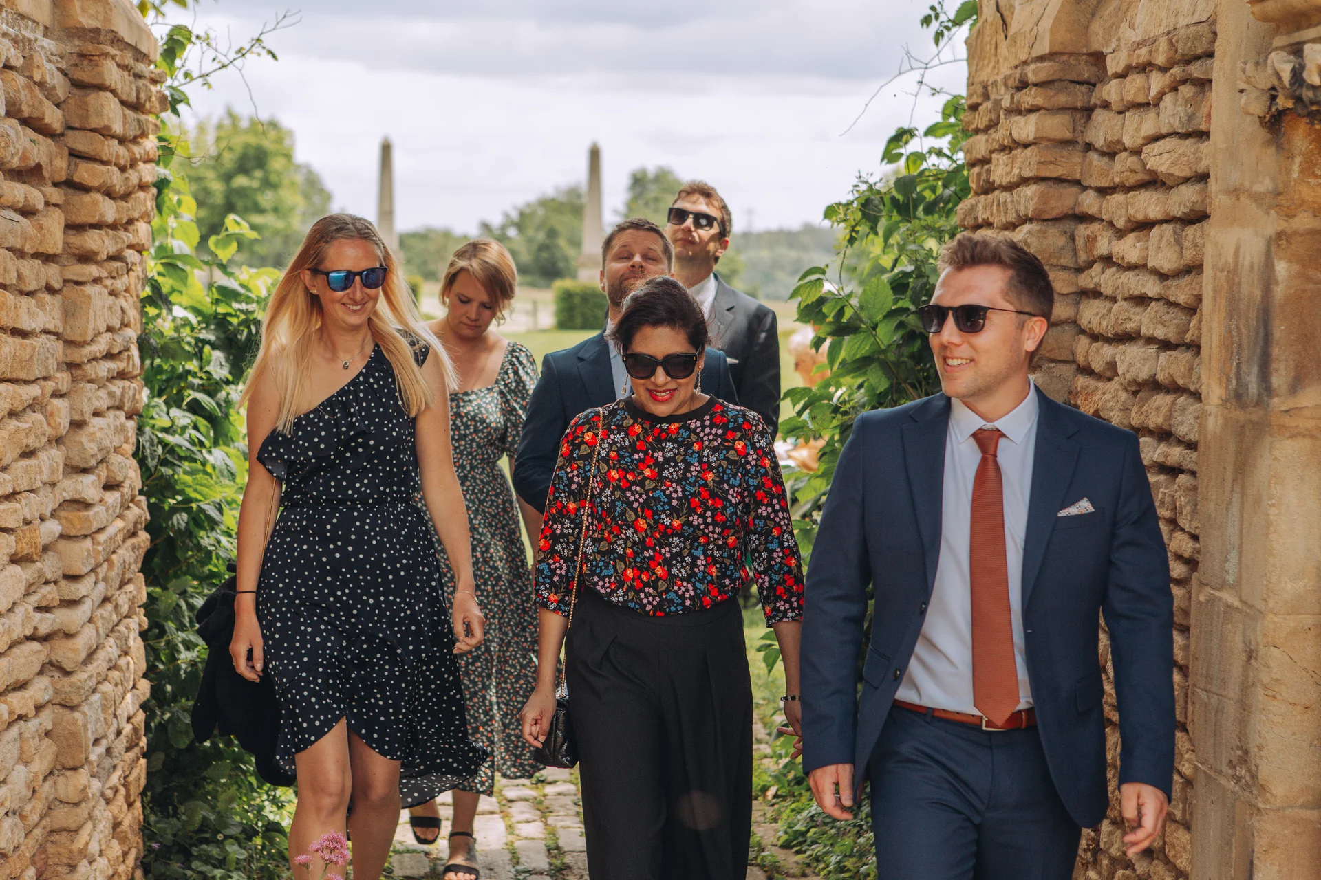A group of well-dressed wedding guests wearing sunglasses walk together outdoors through a historic stone archway during a summer celebration.