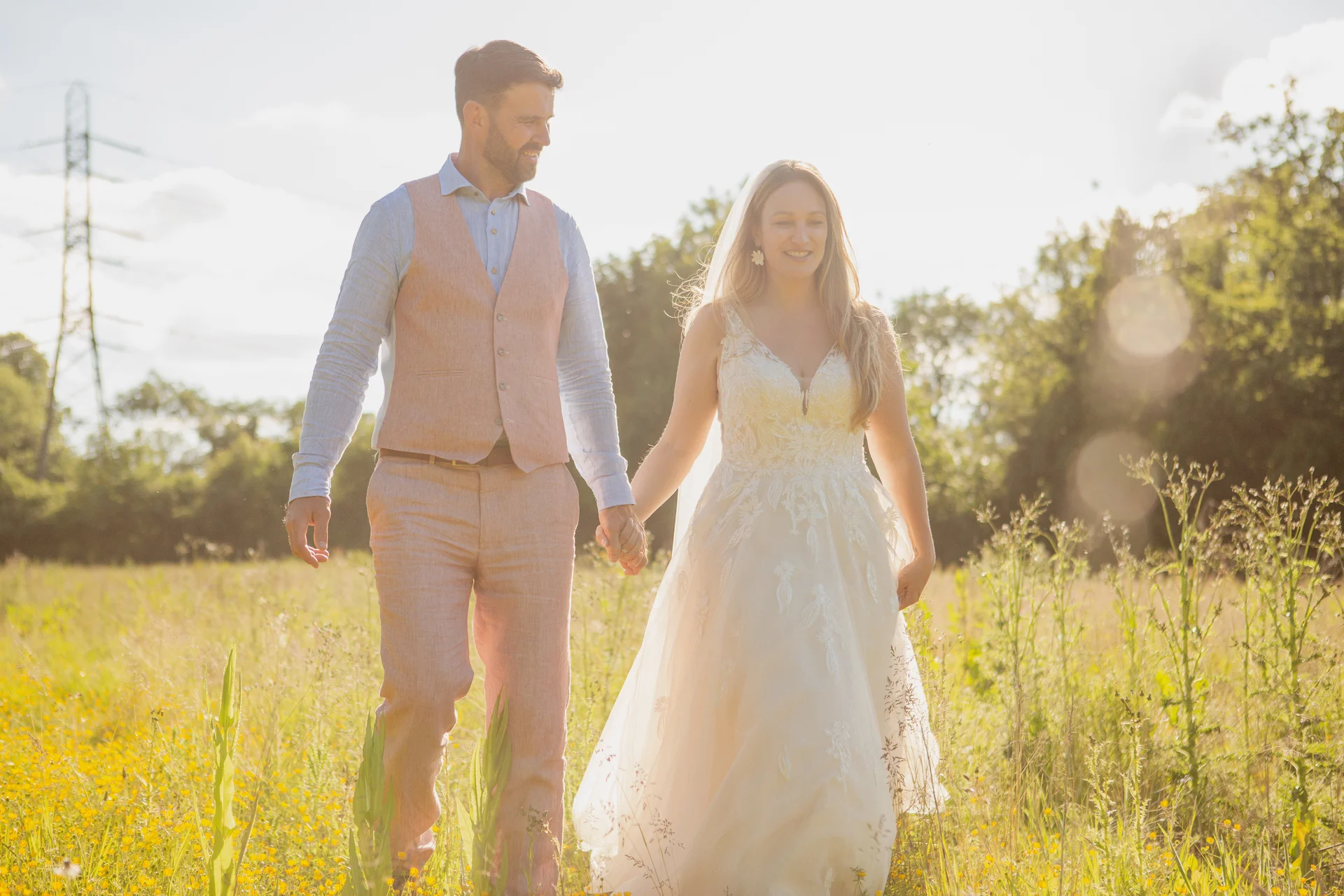 A newlywed couple happily walk hand in hand through a sunlit wildflower meadow during golden hour.