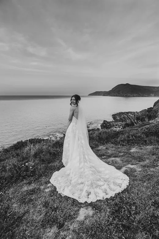 A stunning bride wearing a lace wedding dress with a long train and veil glances back over her shoulder while standing on a dramatic clifftop overlooking the sea.