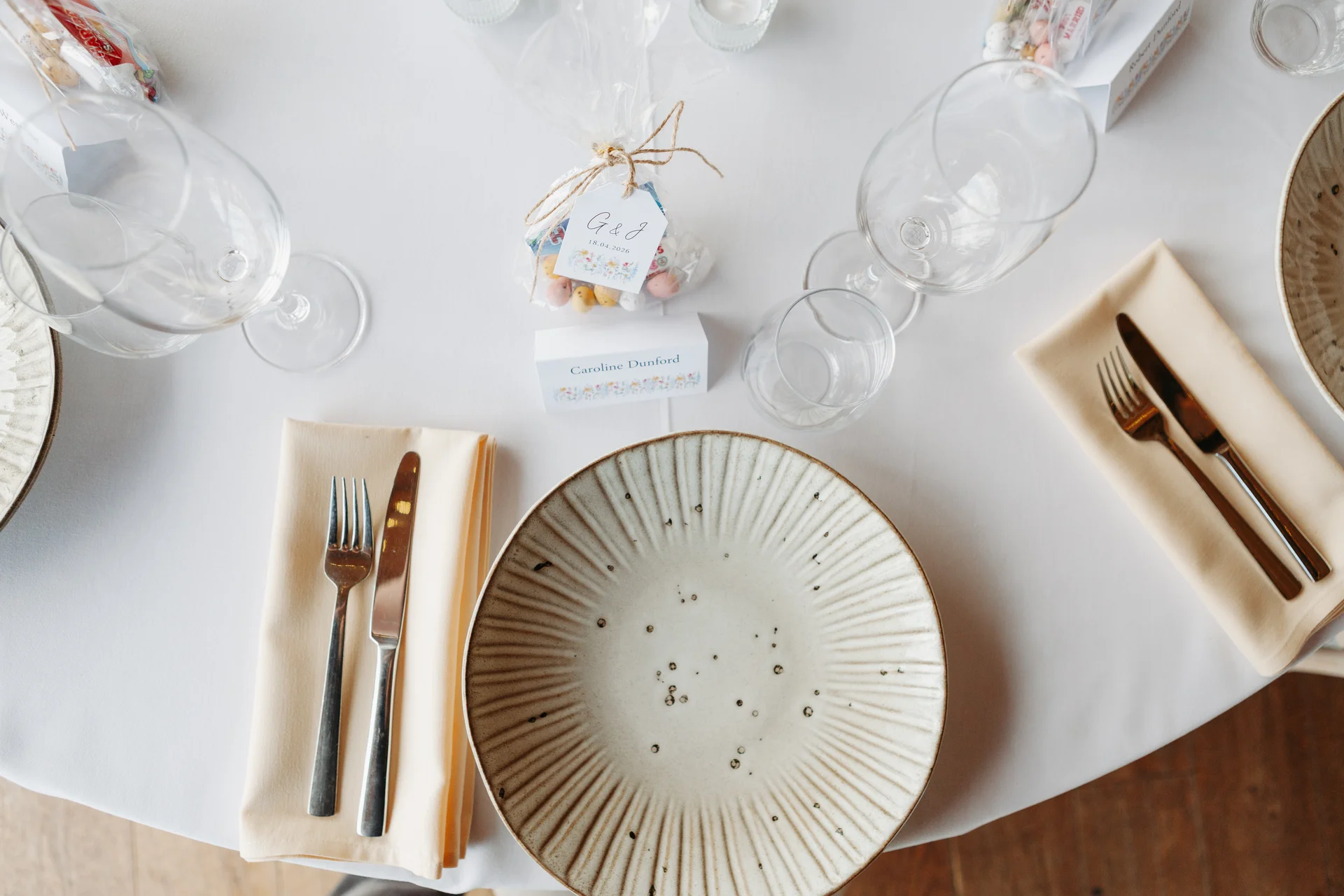 An overhead view of an elegantly set wedding table featuring textured ceramic plates, folded cream napkins, place cards, and sweet favour bags.