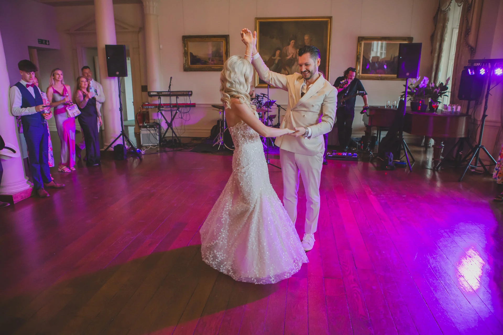 The newlywed couple share their first dance together on a wooden floor in a grand ballroom illuminated by vibrant pink uplighting, with guests and a live band watching in the background.