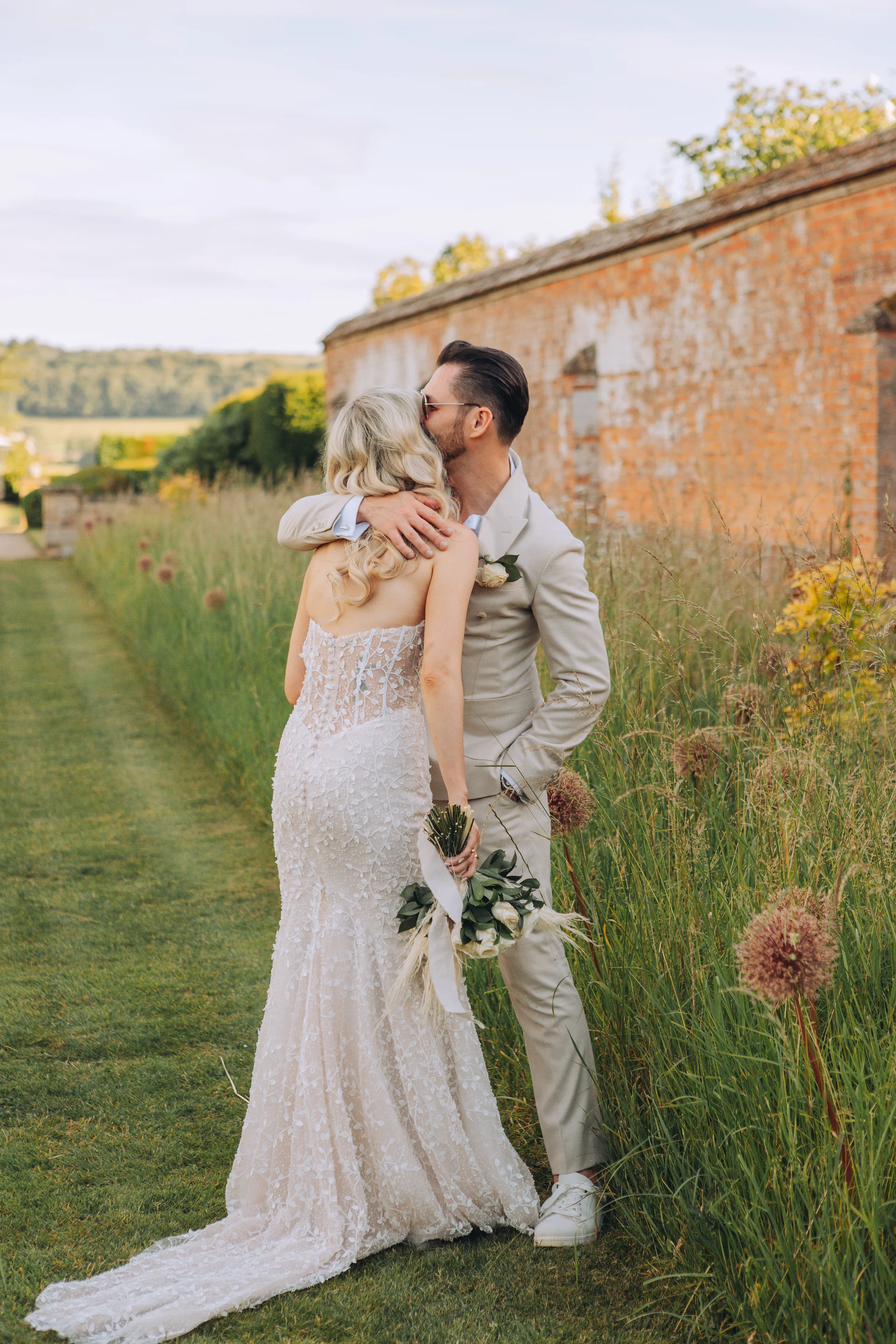 A bride and groom share a tender embrace outdoors next to a rustic brick wall and tall meadow grass during golden hour.