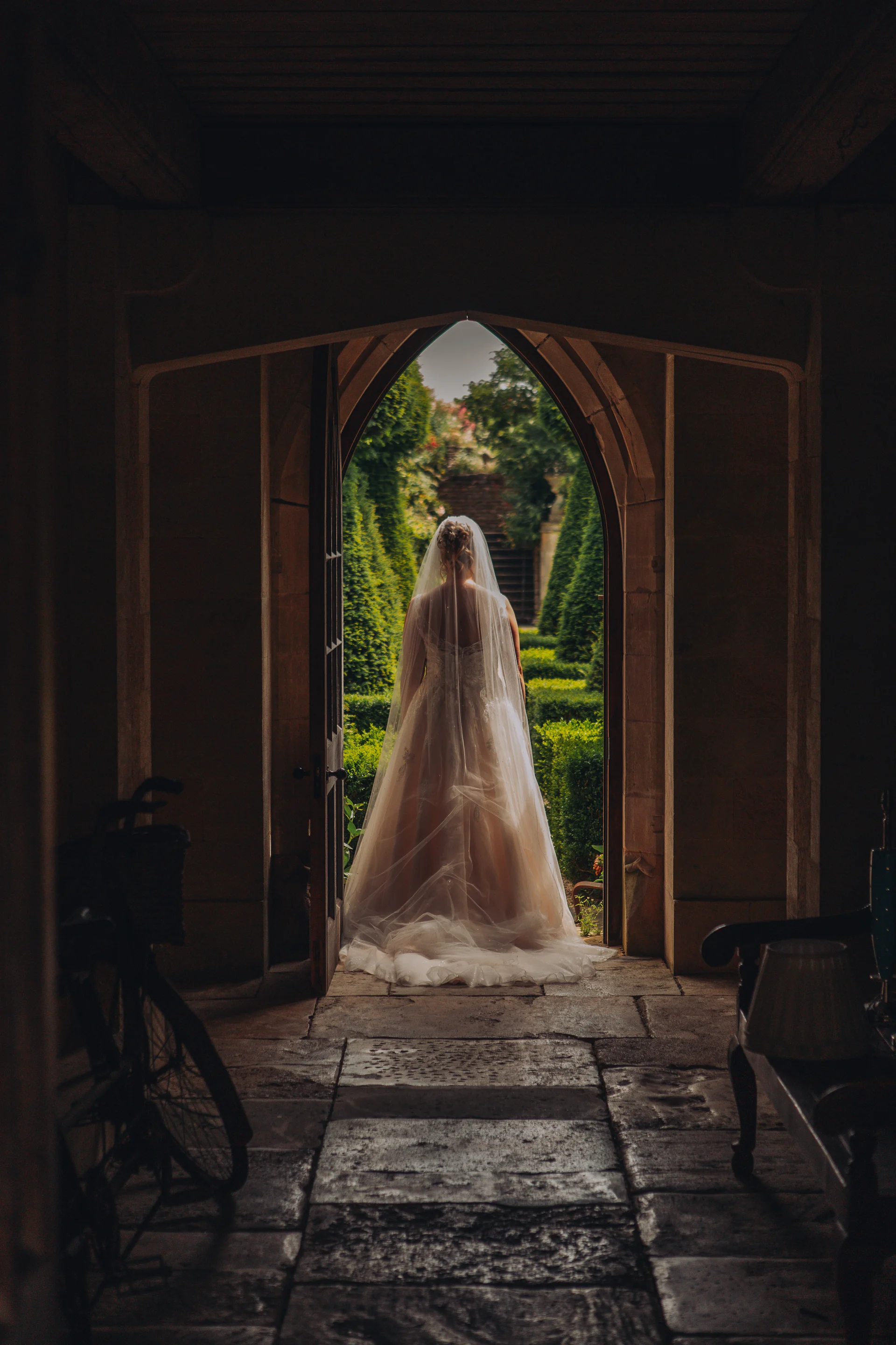 A bride viewed from behind stands framed within a stone archway, looking out towards a lush, manicured garden.