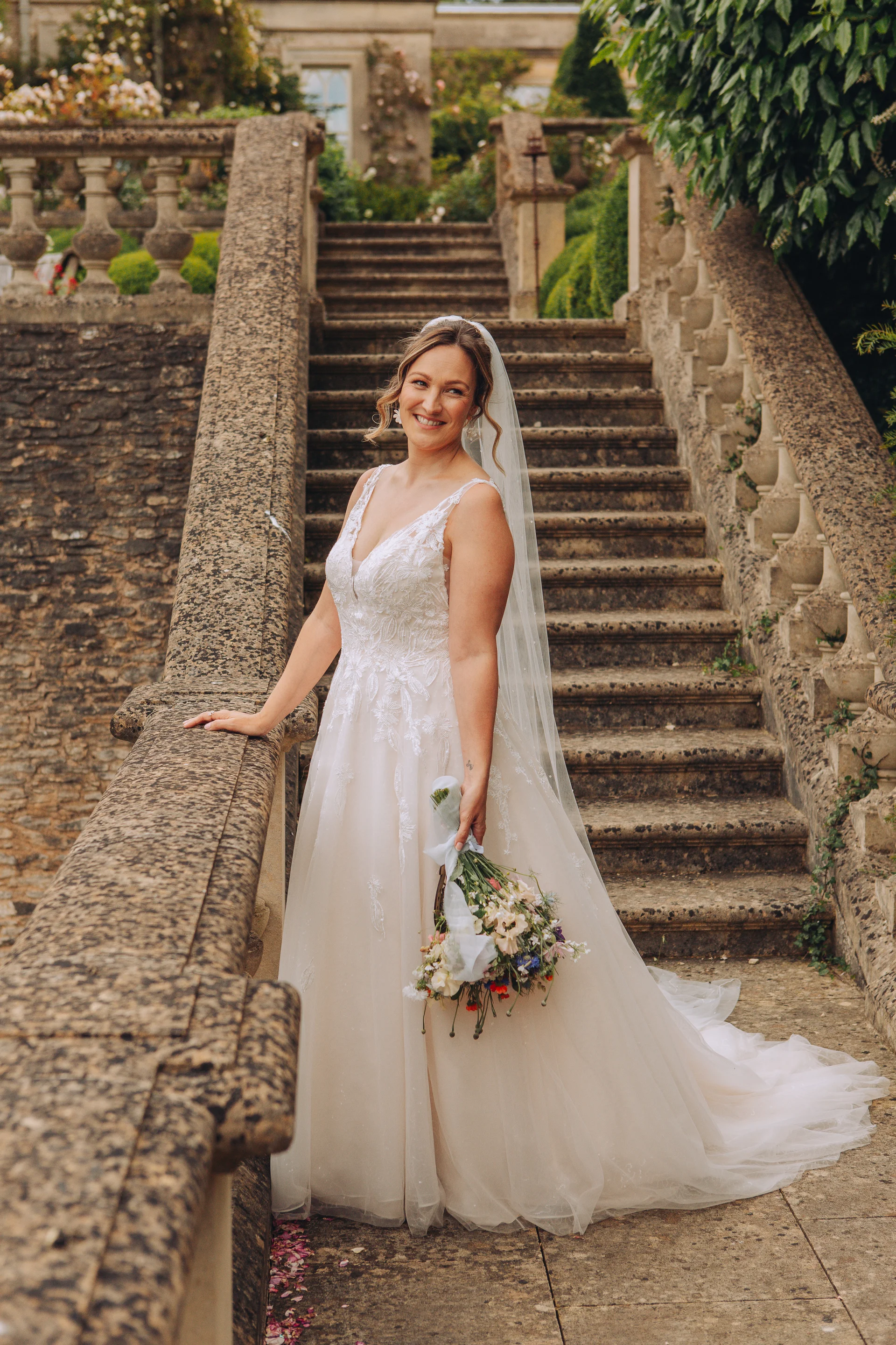 A smiling bride wearing an A-line lace wedding dress and veil stands on a grand stone staircase at a historic manor house, holding a rustic wildflower bouquet.