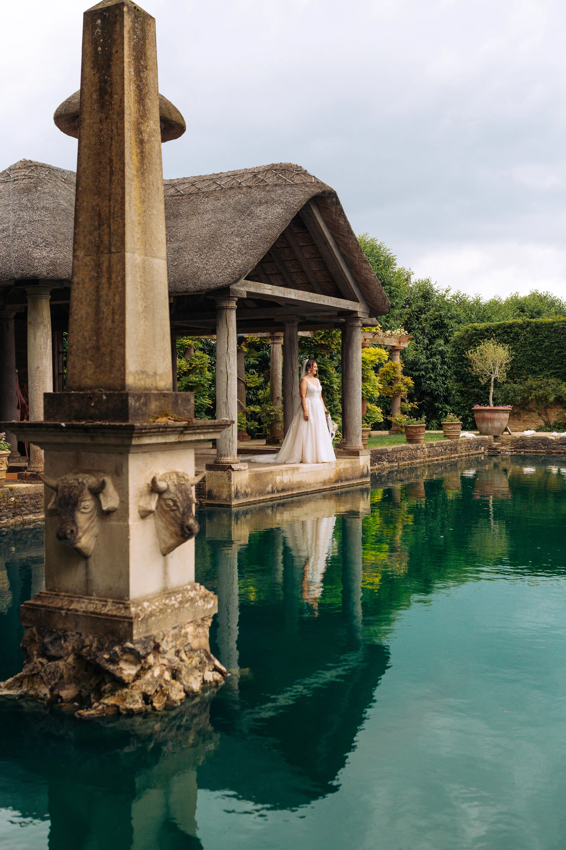 A solo bride in an a line gown stands gracefully by an ornamental lake reflecting her image near a historic stone pavilion.