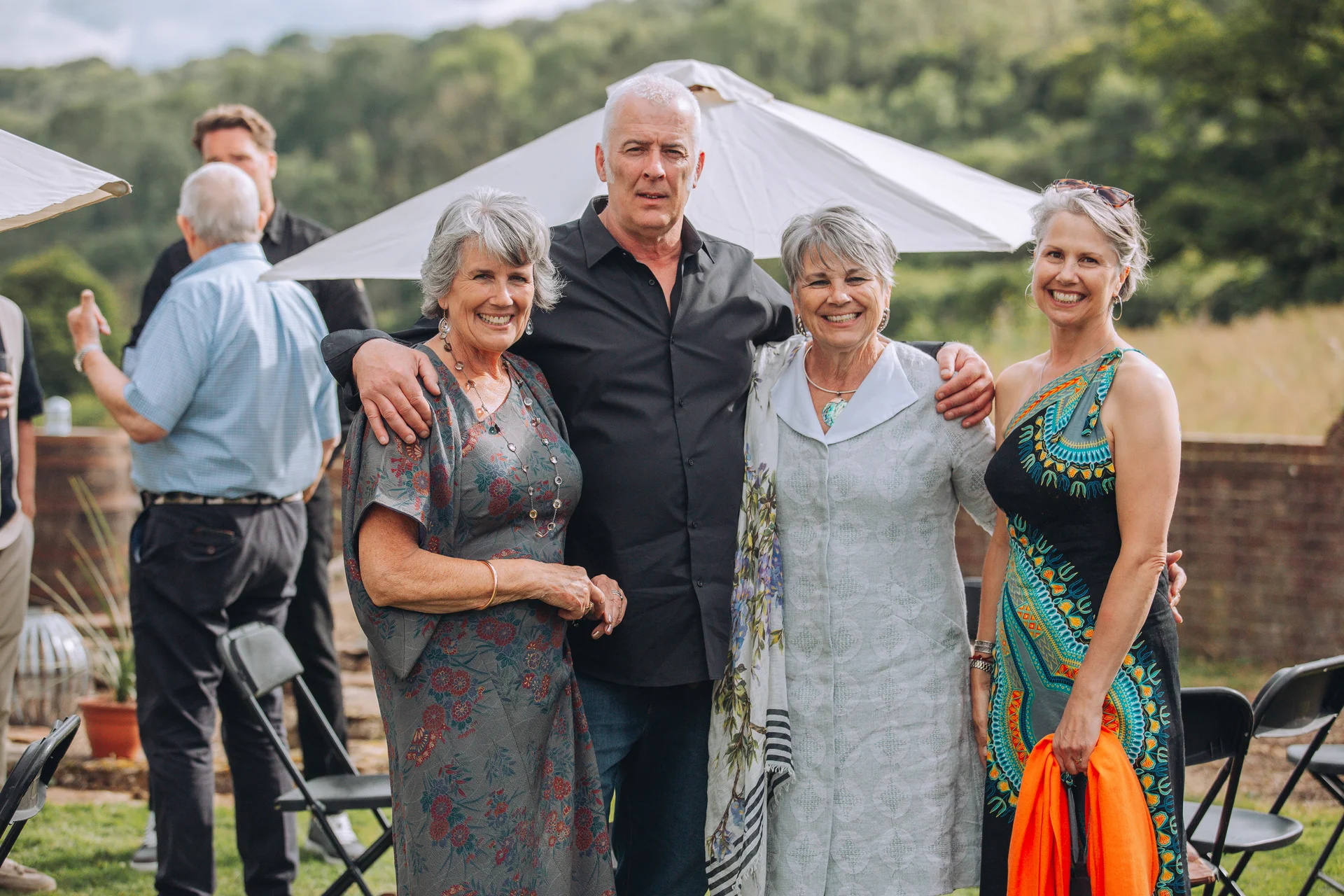A group of older wedding guests smile happily outdoors in a garden setting beneath white parasols.