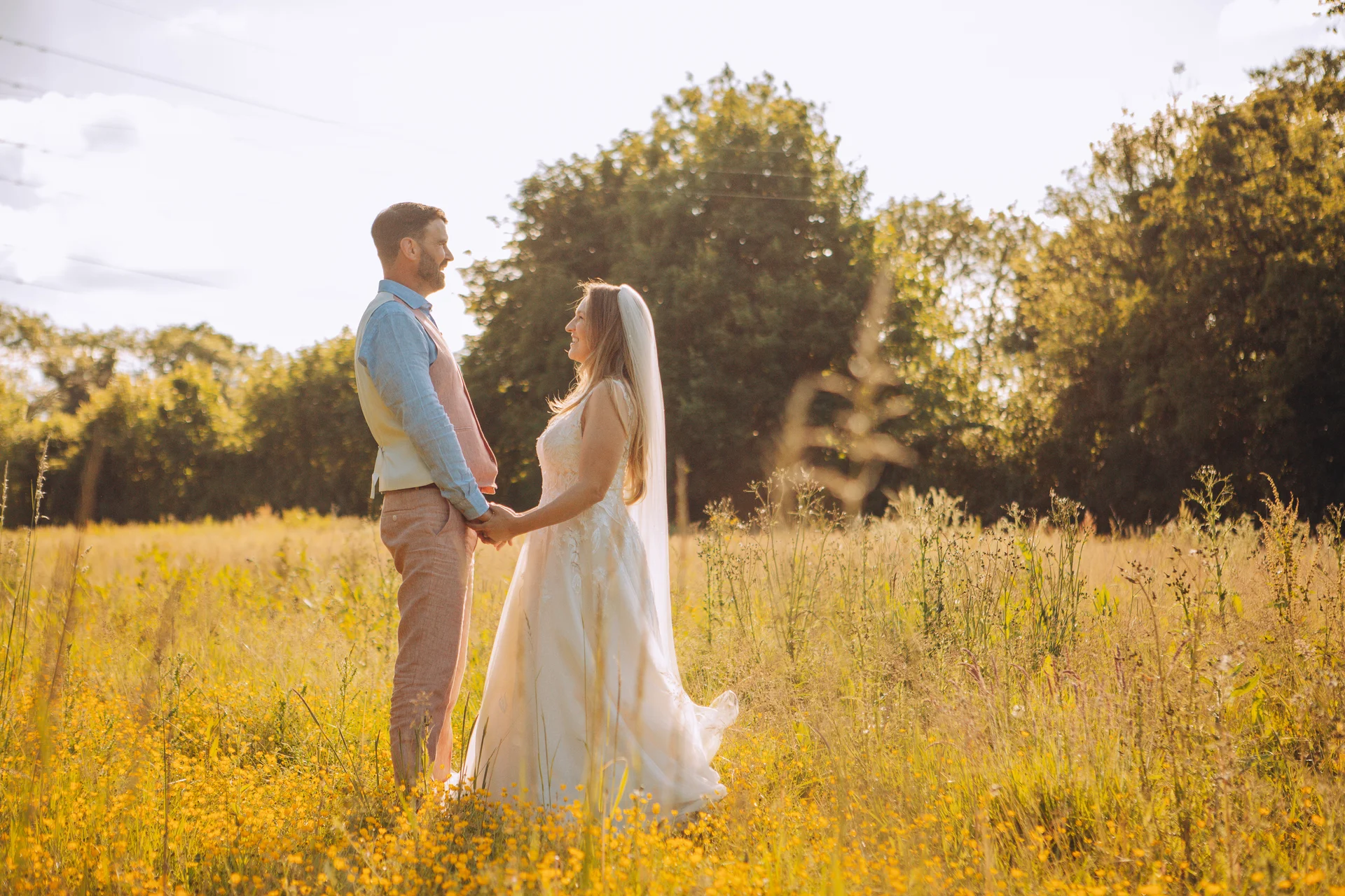 Newlywed bride and groom hold hands and gaze at each other in a sunlit wildflower meadow during golden hour.