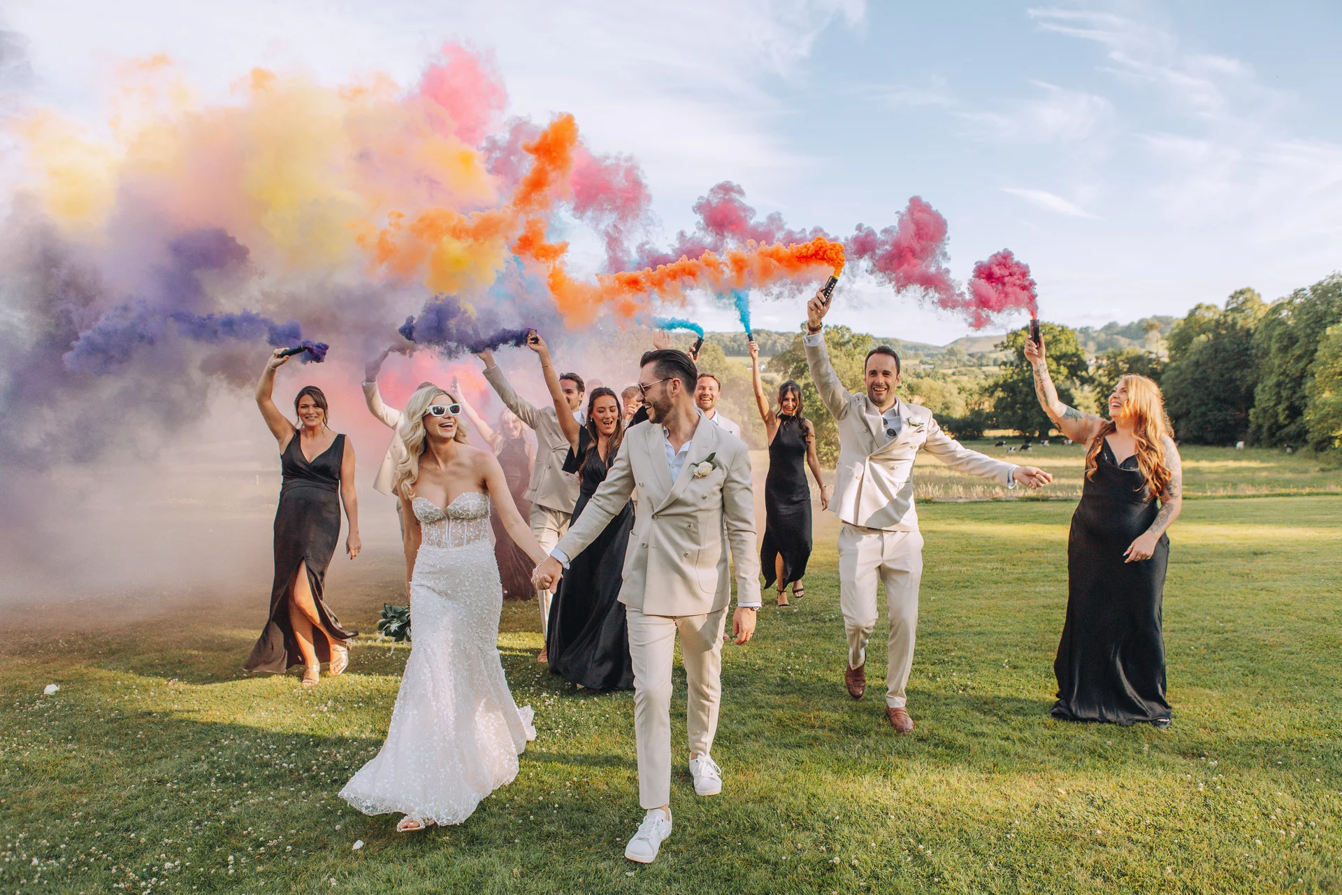 A bride and groom walk hand in hand across a grassy field while their wedding party trails behind them, holding colourful smoke grenades that fill the sky with a vibrant cloud of hues.