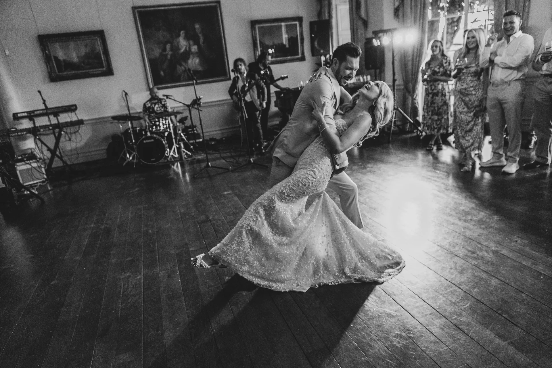 The newlyweds share an enthusiastic dip during their first dance on a wooden ballroom floor while guests watch and a band plays in the background.