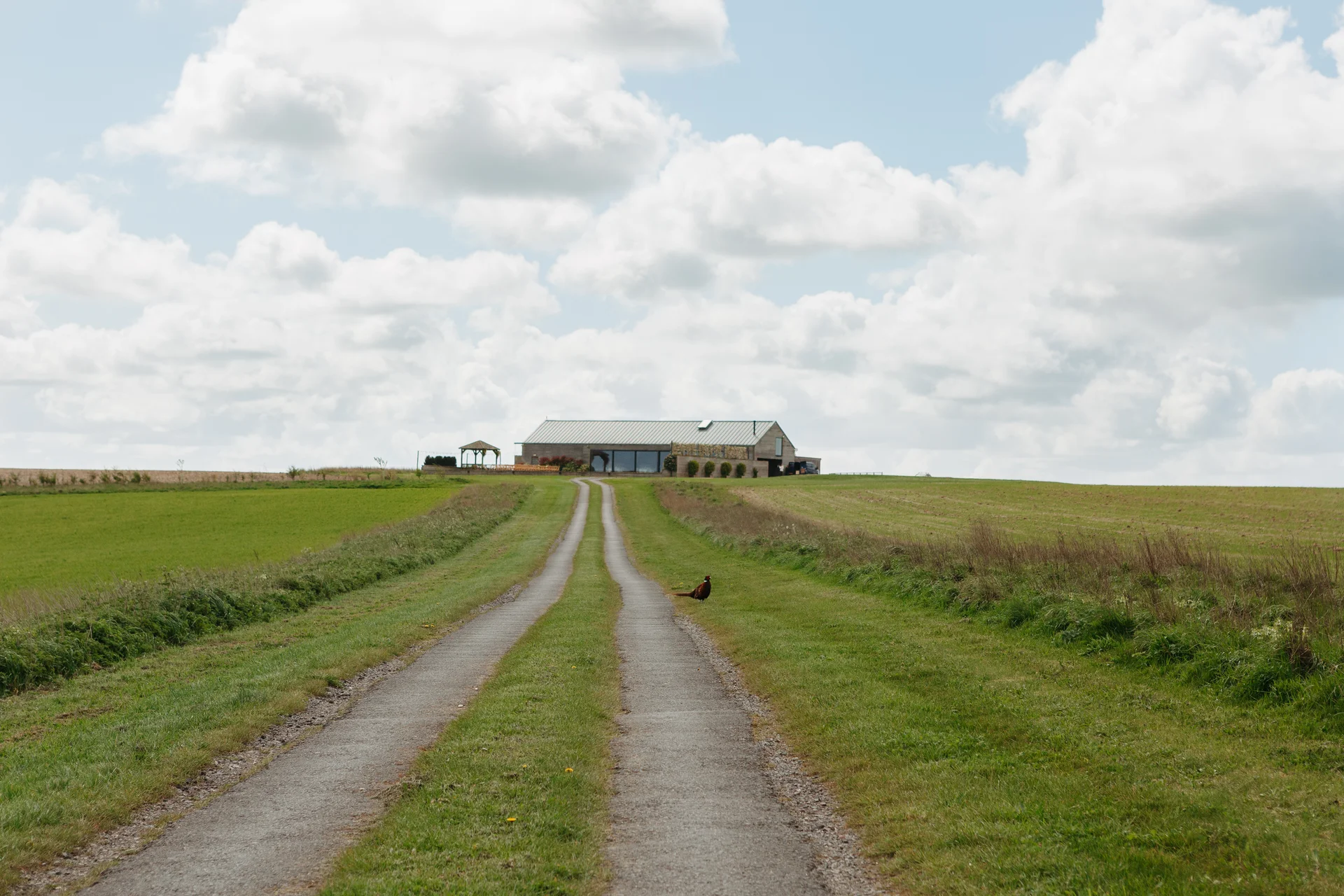 A long, narrow country track leads up a gentle hill toward a modern converted barn wedding venue set amidst expansive green fields and a cloudy blue sky.