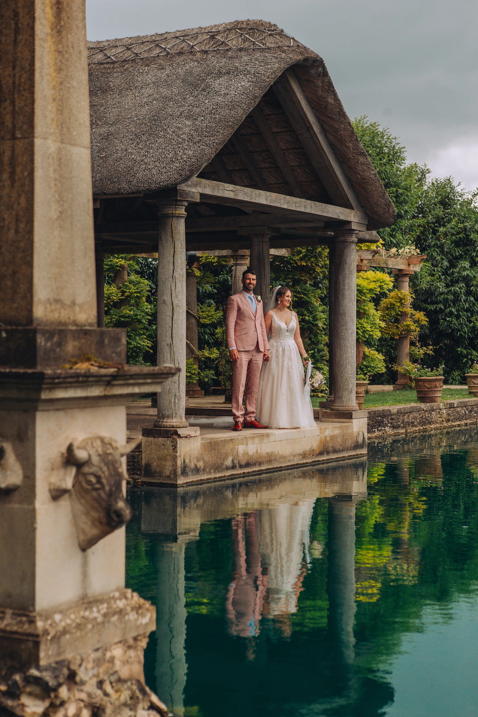 A newlywed couple stands together holding hands beneath a thatched pavilion by a reflective lake on their wedding day.