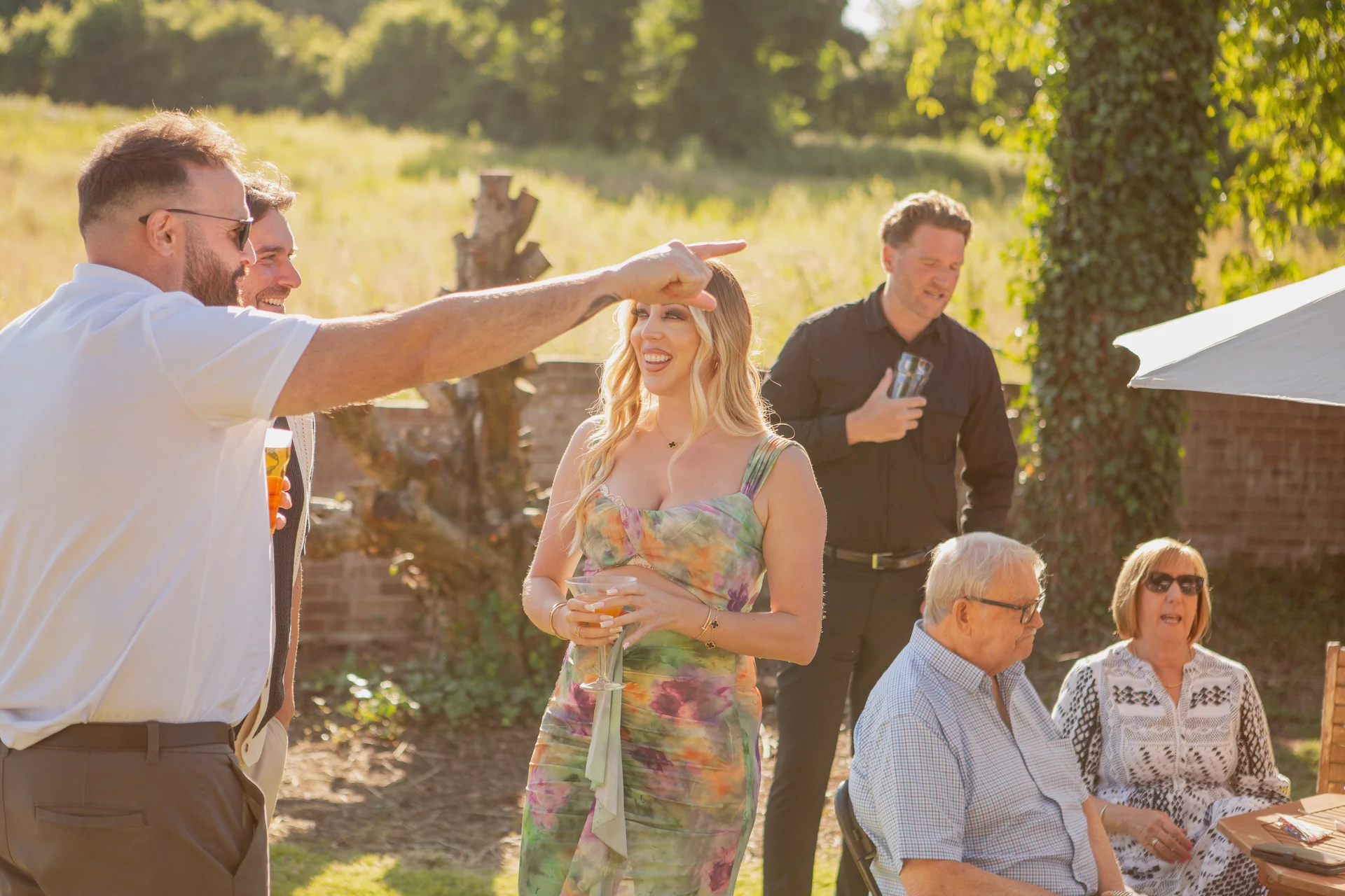 Wedding guests laugh and mingle outdoors during a sunlit drinks reception in a scenic garden setting.