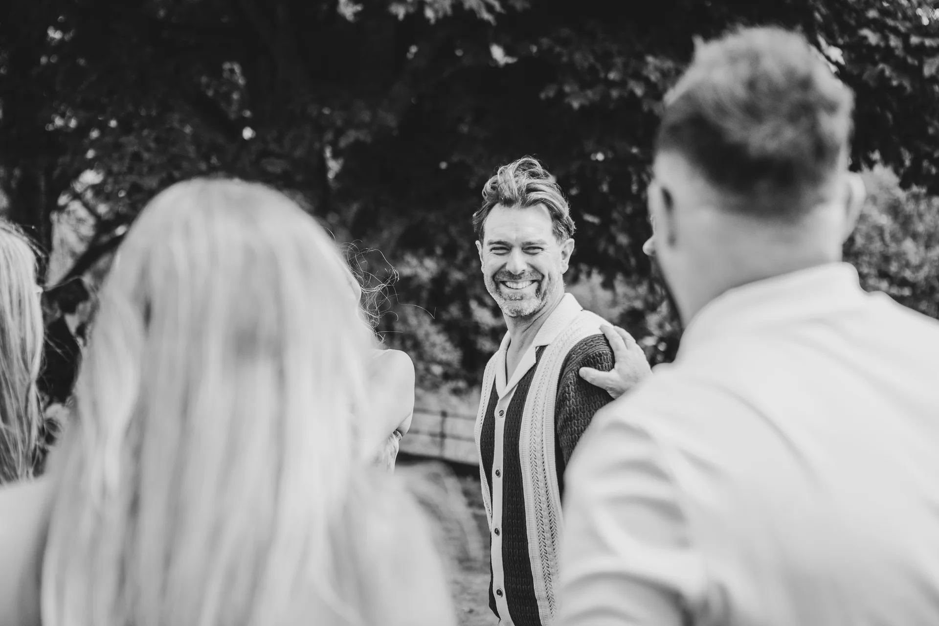 A smiling man looks back over his shoulder amongst a gathering of wedding guests in a candid black and white shot.