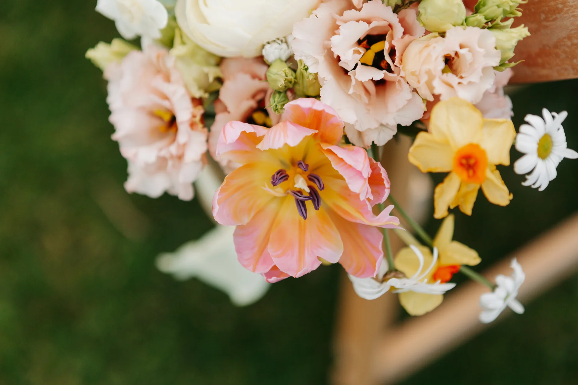 A close up view of a colourful spring wedding bouquet featuring peach and pink parrot tulips, yellow daffodils, and white daisies against a soft blurred background.