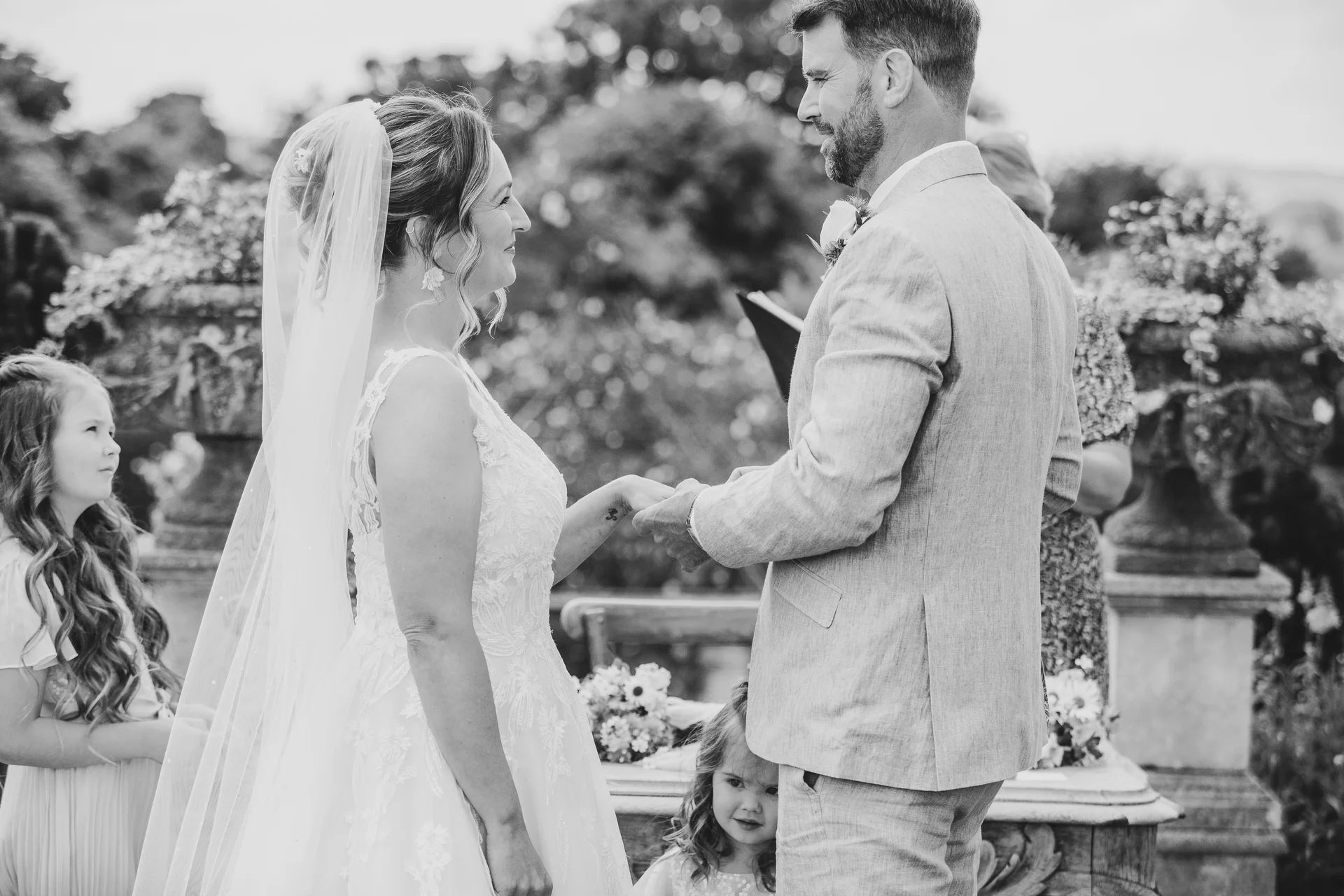 An emotional bride and groom exchange rings during an outdoor wedding ceremony while flower girls look on in a garden setting.