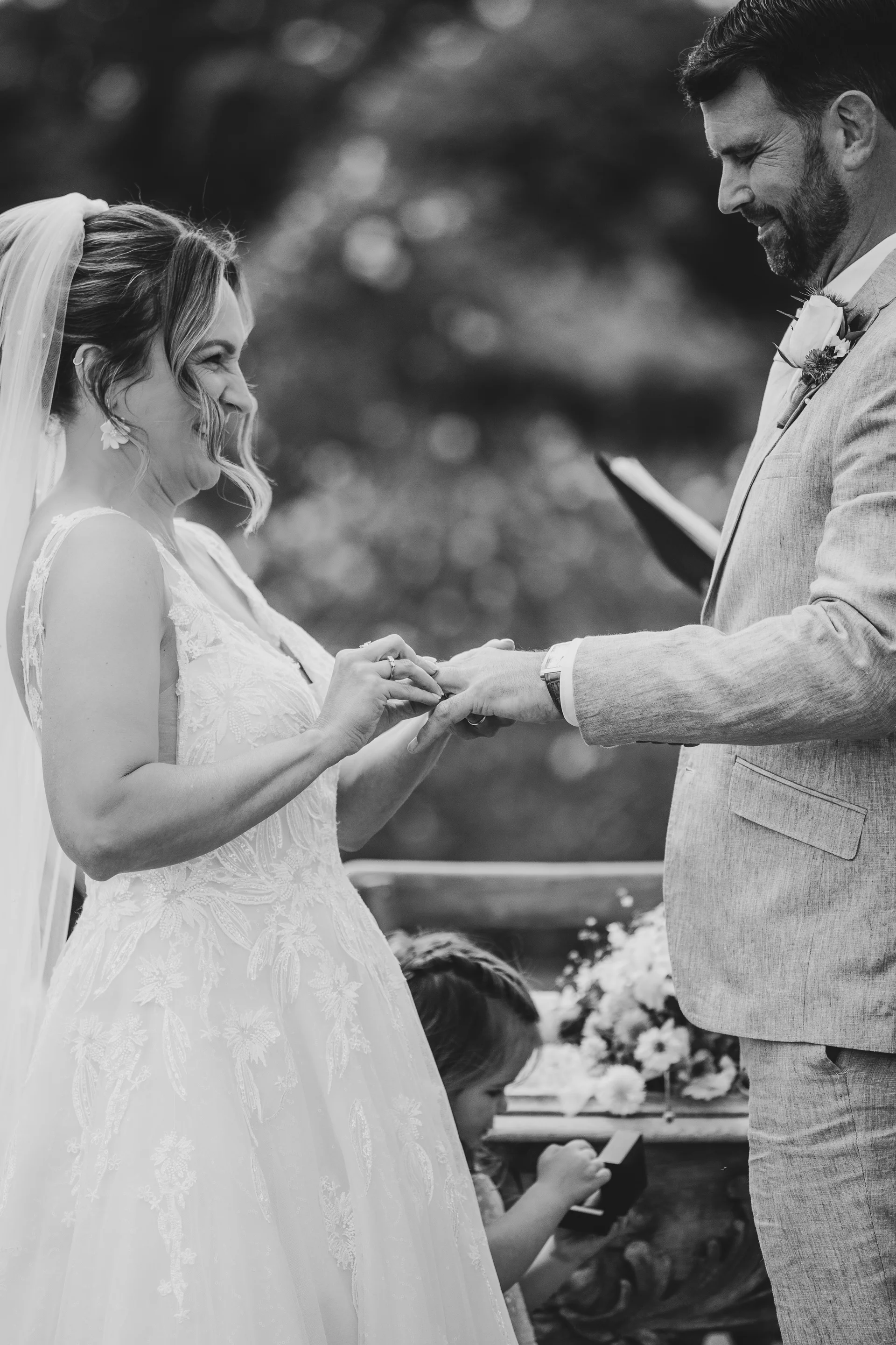 A joyful bride and groom exchange rings during an outdoor ceremony, sharing a laugh while the bride places the band on his finger.