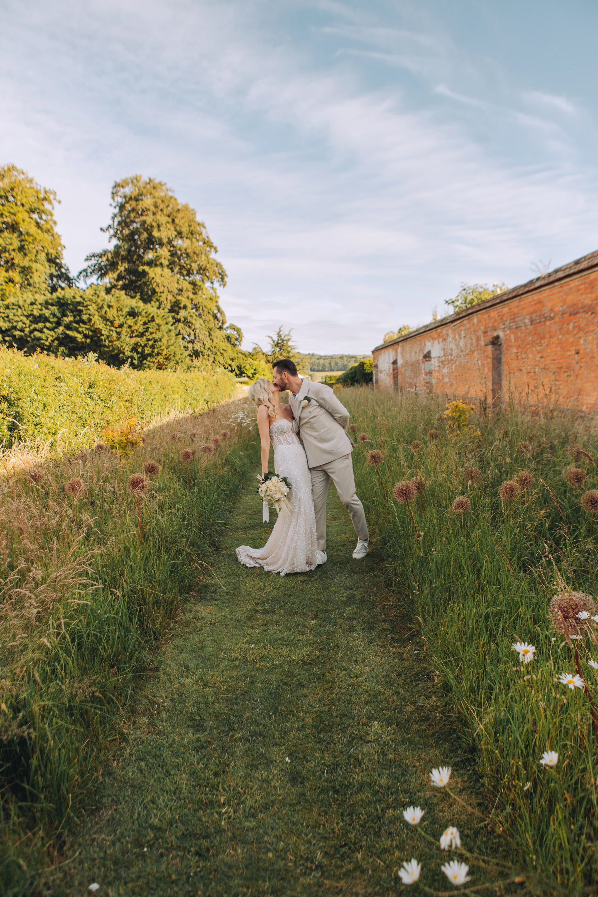 A newlywed couple shares a romantic dip kiss on a grassy path framed by tall wildflowers and an old brick wall during golden hour.