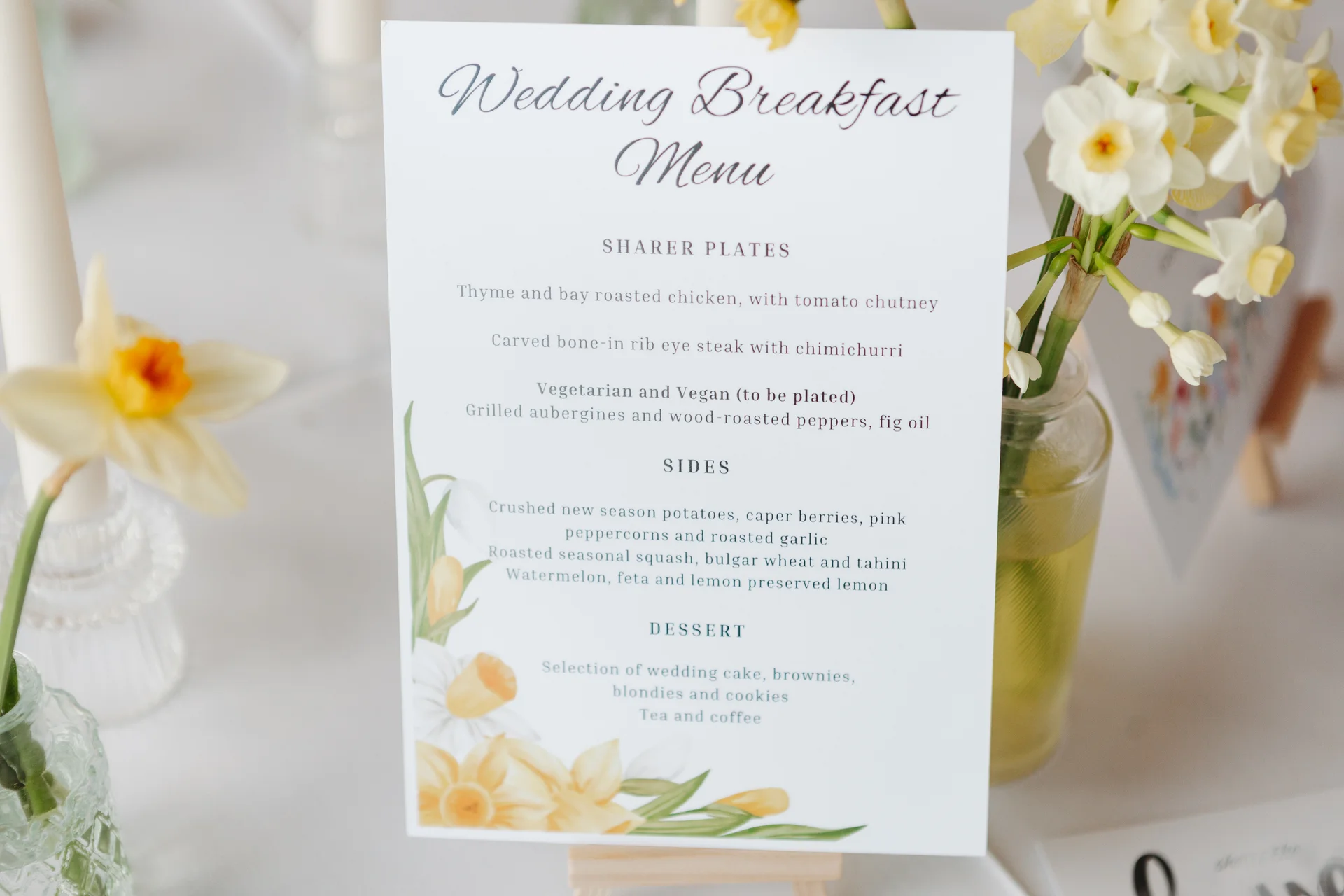 A close-up of a wedding breakfast menu card set amidst delicate yellow daffodils and glass bud vases on a reception table.