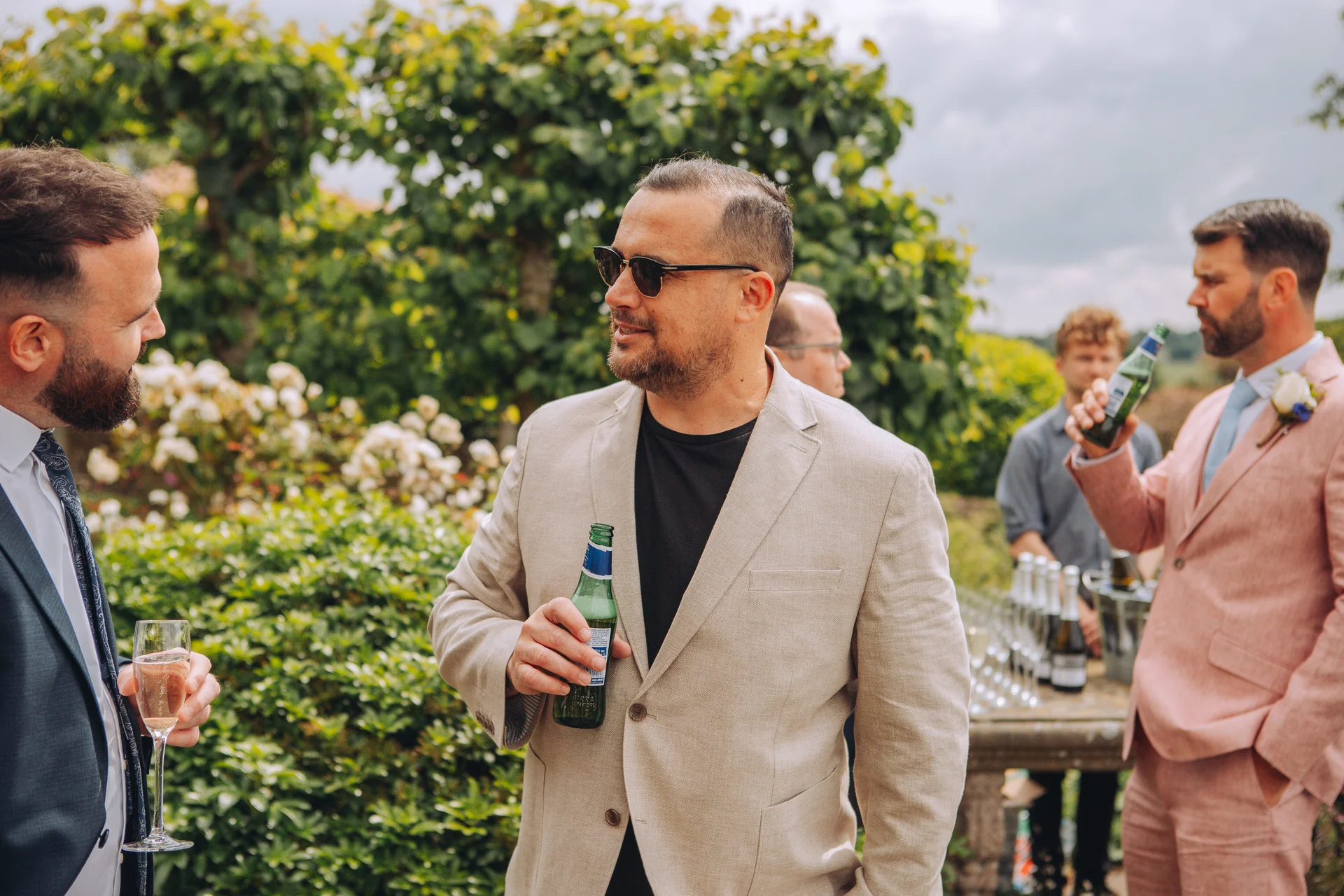 Wedding guests enjoy drinks and conversation outdoors during a relaxed garden reception on a sunny summer day.
