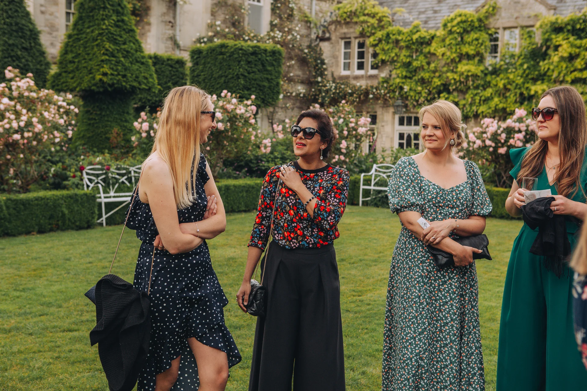 Four female wedding guests dressed in stylish patterned dresses and sunglasses chat and mingle on a manicured lawn outside a historic stone manor house.