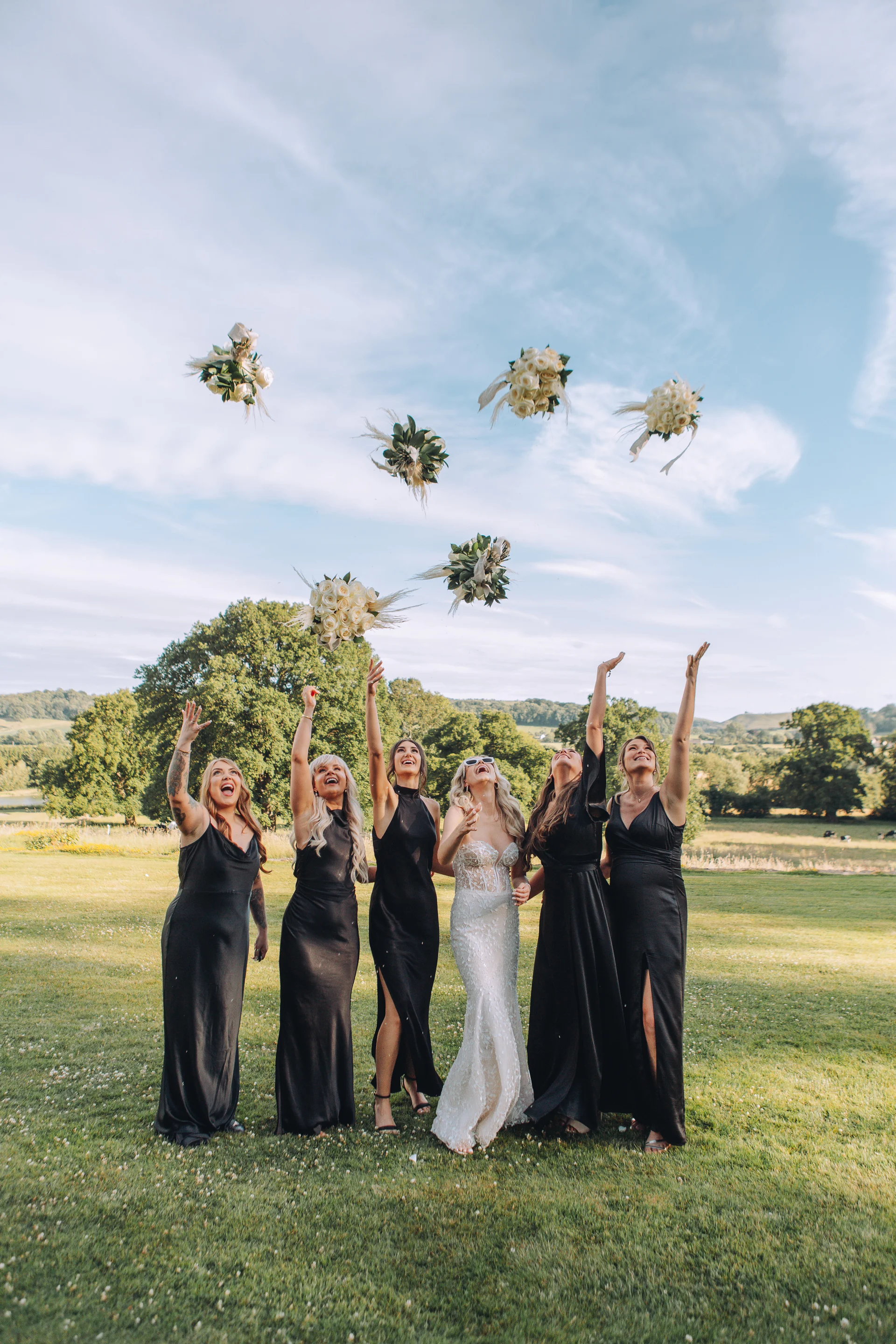 A joyful bride and her bridesmaids in black dresses enthusiastically toss their floral bouquets into the air outdoors in a lush green field.
