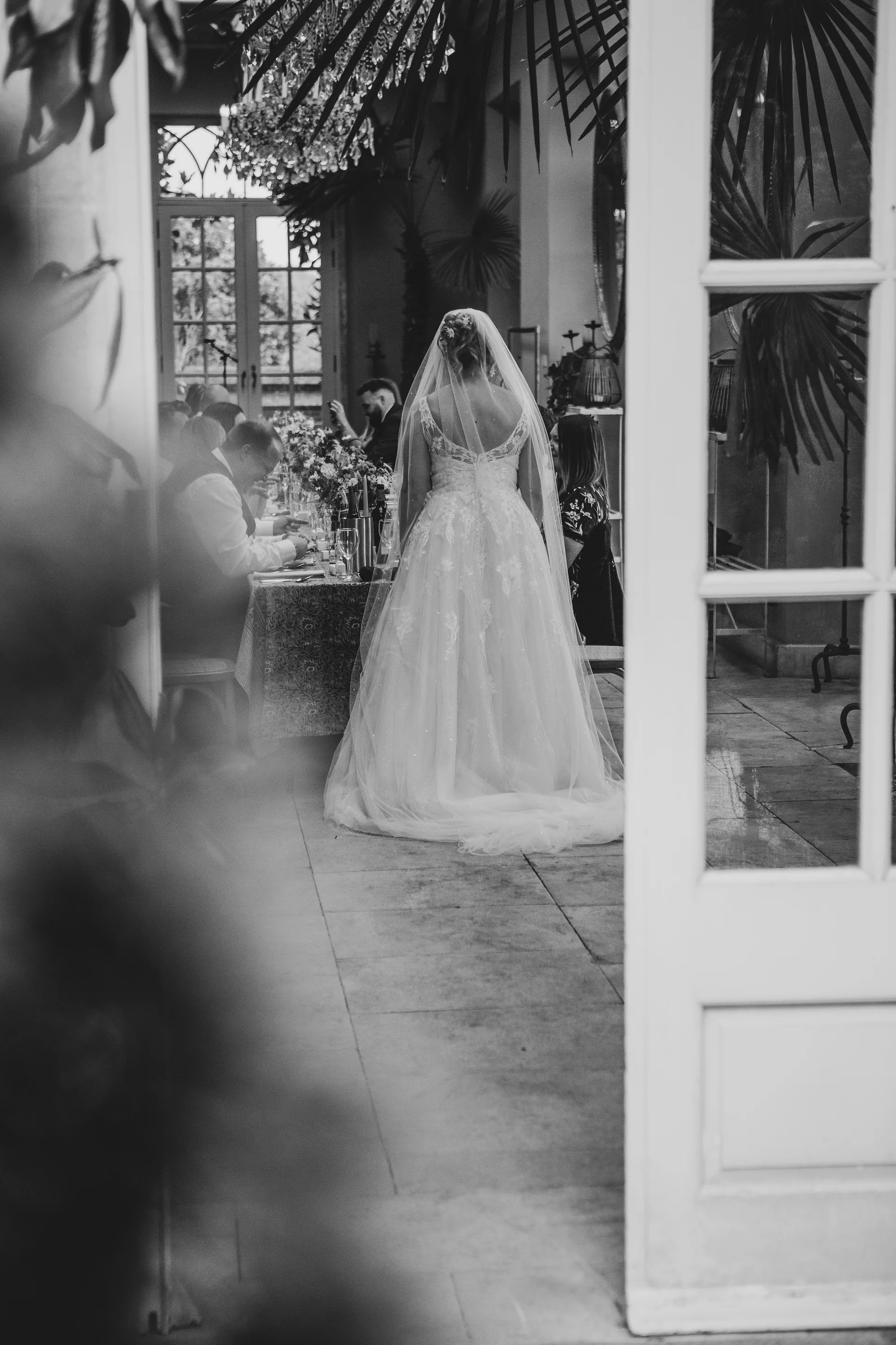 Captured in black and white through an open doorway, a bride in a lace gown and chapel veil stands looking towards guests seated at a long banquet table inside a light-filled conservatory.