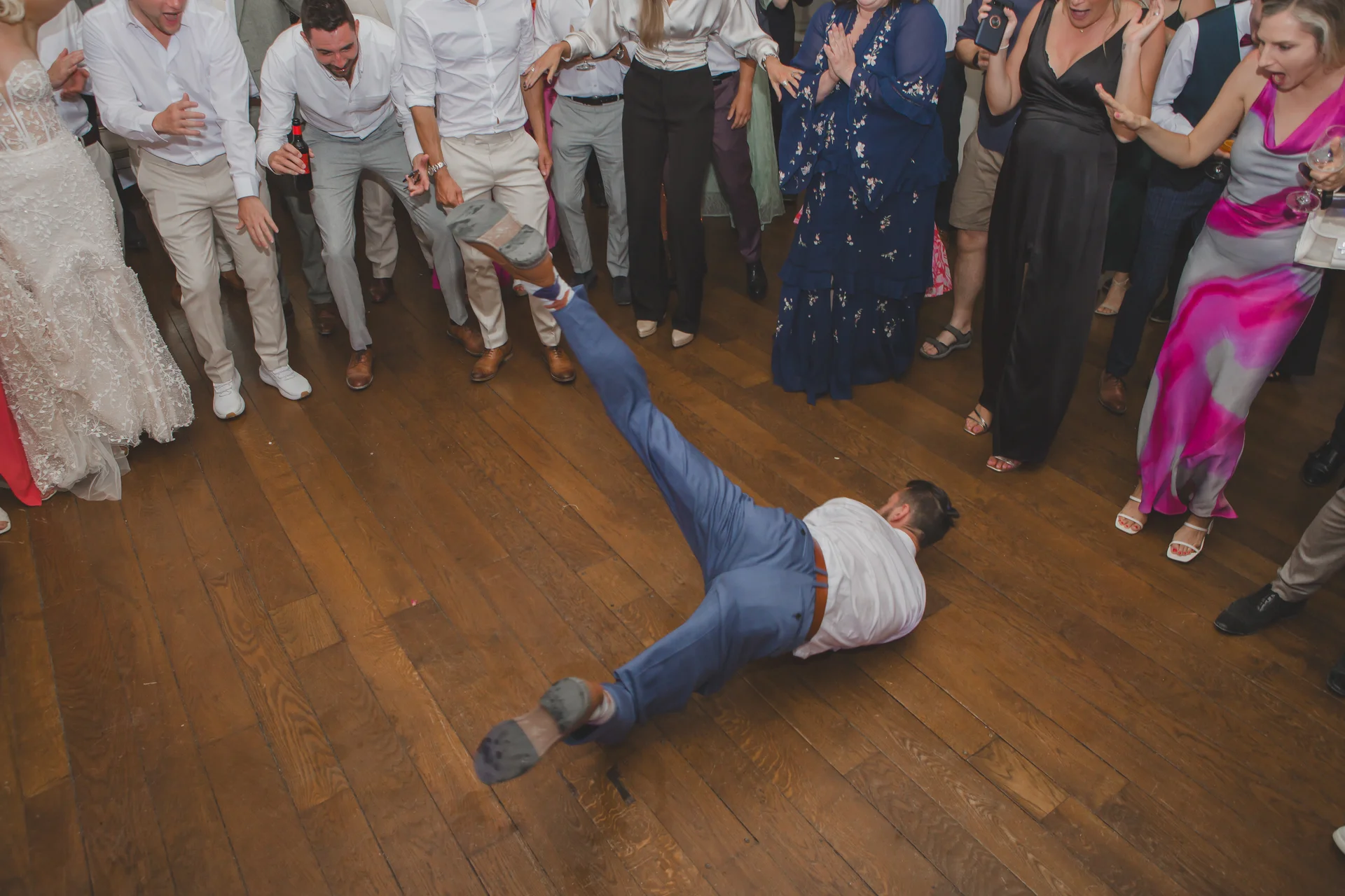 A guest slides across the wooden dance floor in a blue suit while surrounding wedding guests cheer and laugh.