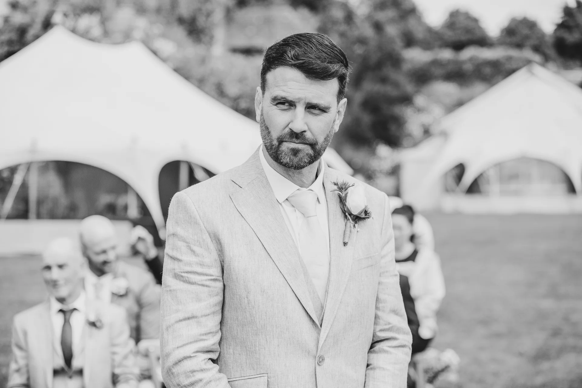 A groom in a light grey suit with a boutonniere looks away with a thoughtful expression during an outdoor marquee wedding ceremony.