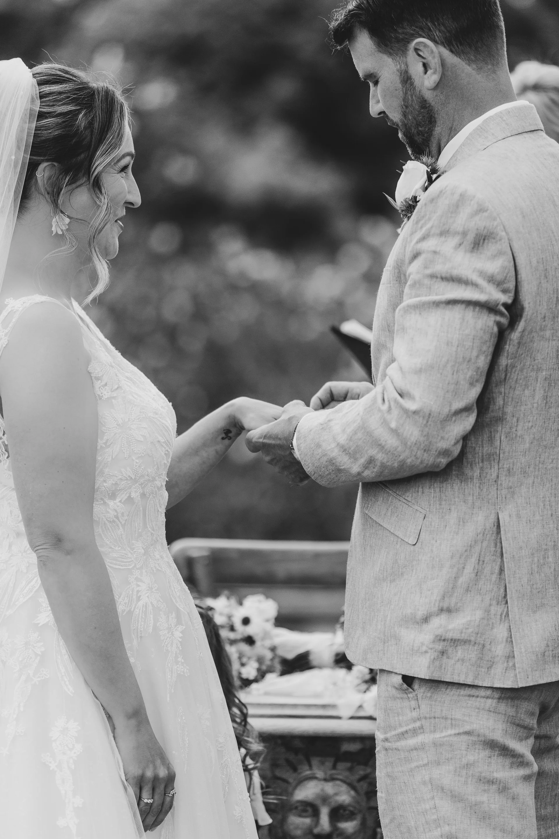 A newlywed couple exchange rings during an outdoor ceremony, with the groom gently placing the band on the bride's finger.
